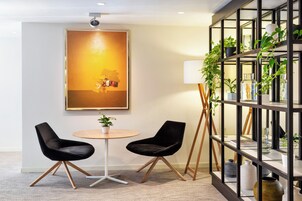 Ein modernes Zimmer mit zwei schwarzen Stühlen, einem runden Tisch, einem großen gerahmten Bild, einem Bücherregal mit Pflanzen und einer Stehlampe.