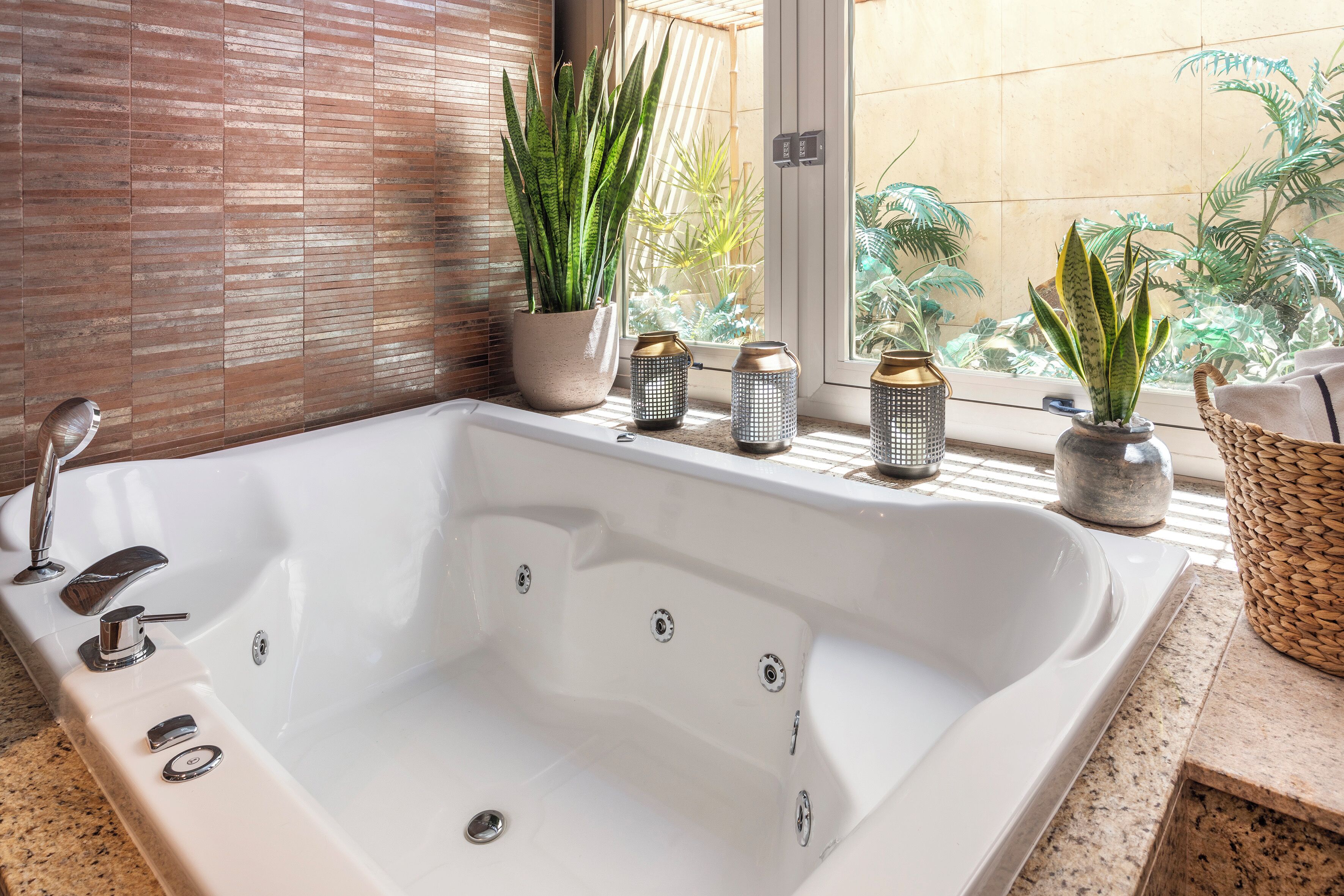 Una bañera blanca grande con múltiples grifos, una planta en maceta y dos faroles decorativos en un alféizar.