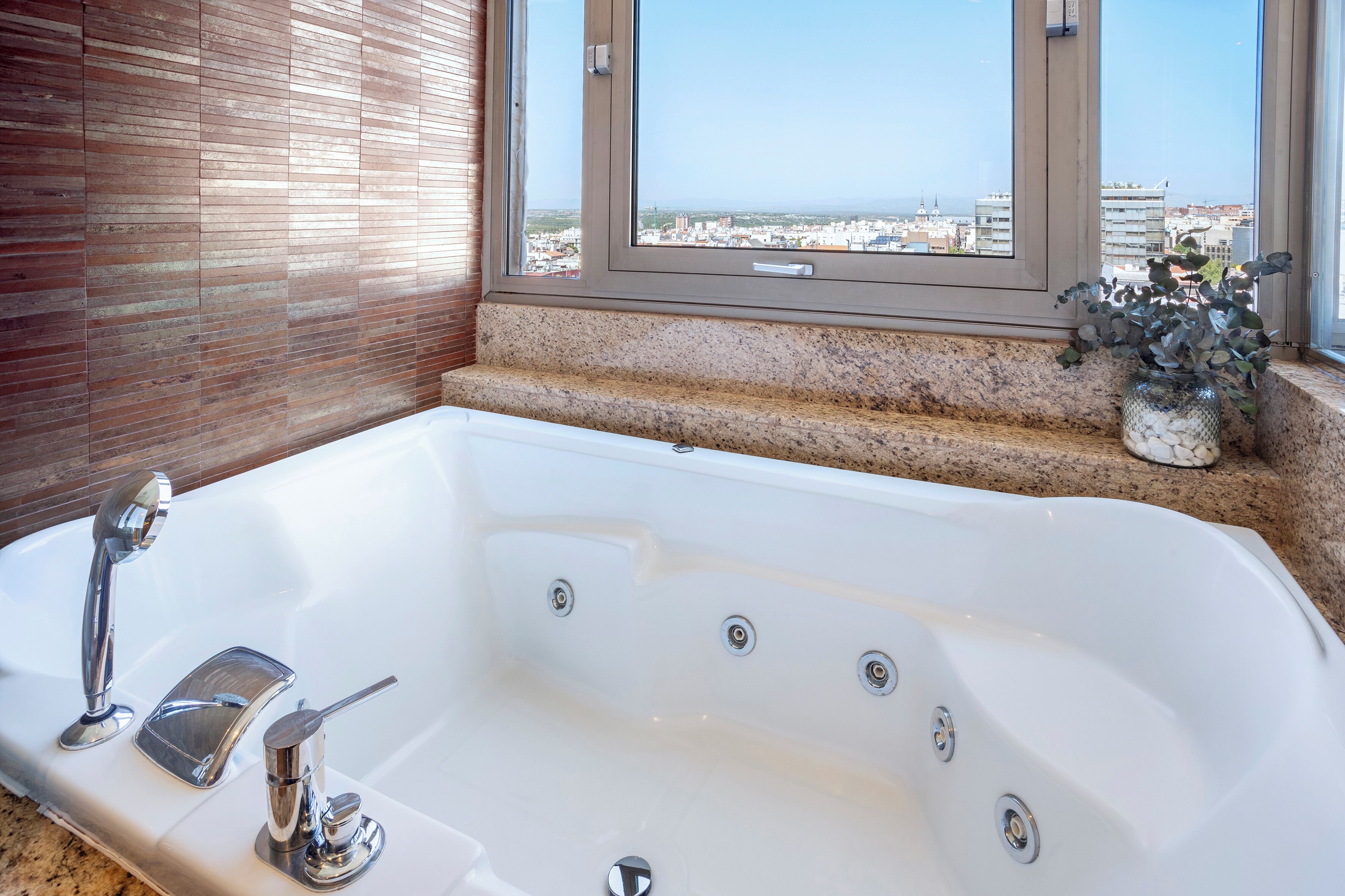 A bathroom with a large bathtub, a window with a view of a cityscape, and a potted plant on the windowsill.