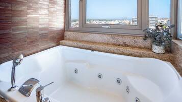 A bathroom with a large bathtub, a window with a view of a cityscape, and a potted plant on the windowsill.