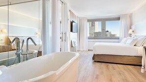 A modern hotel room with a large bathtub, a bed with white linens, a television mounted on the wall, and a view of the city through large windows.