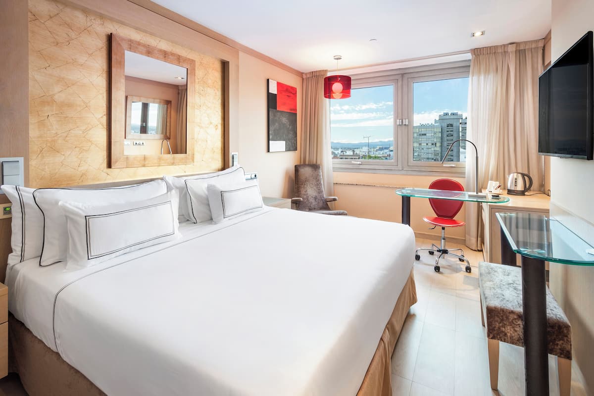 A hotel room with a large bed, a desk with a glass top, a chair, a TV, and a view of the city.