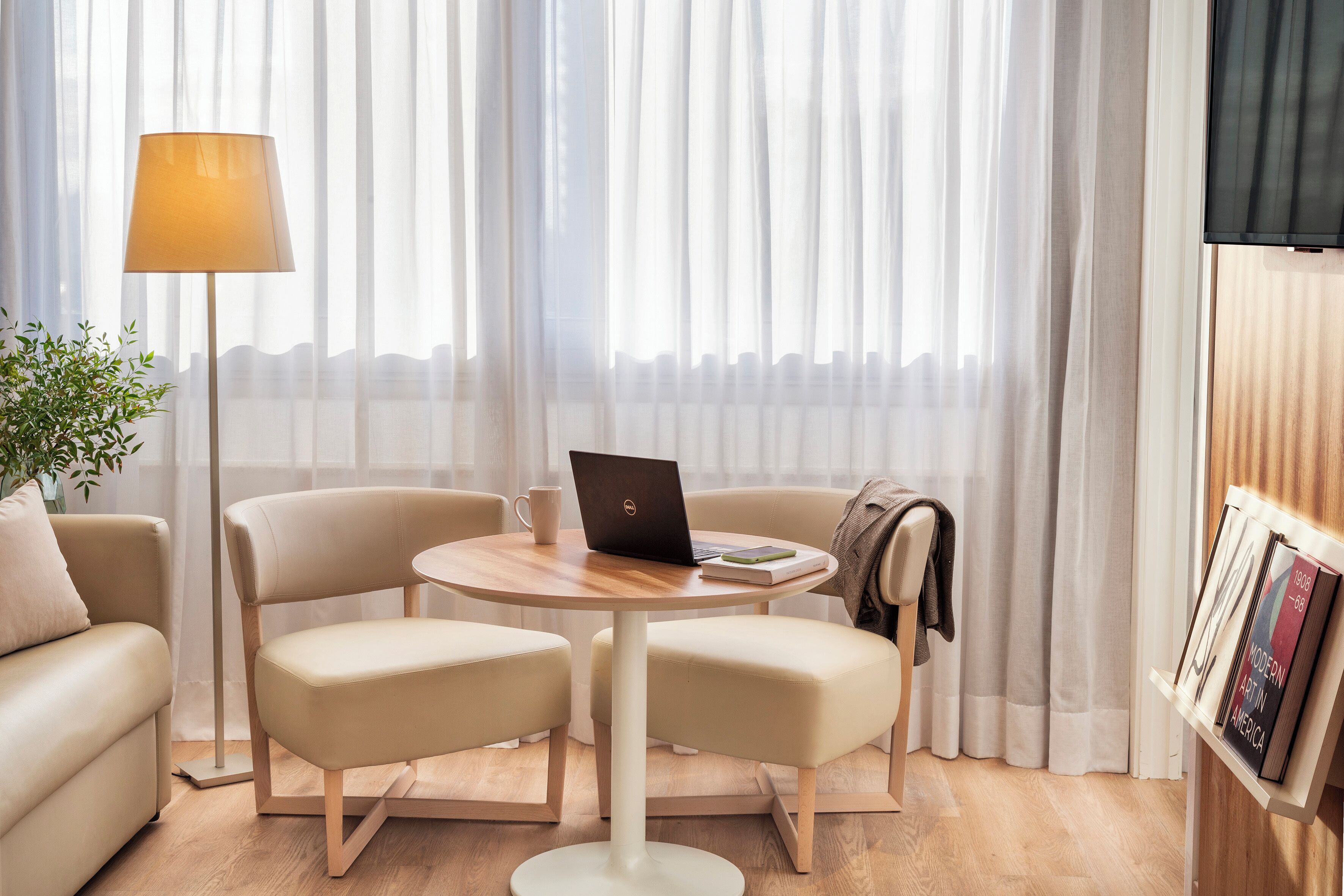 Una sala de estar moderna con una mesa redonda de madera, dos sillones color beige, una computadora portátil, una lámpara y una estantería llena de libros.