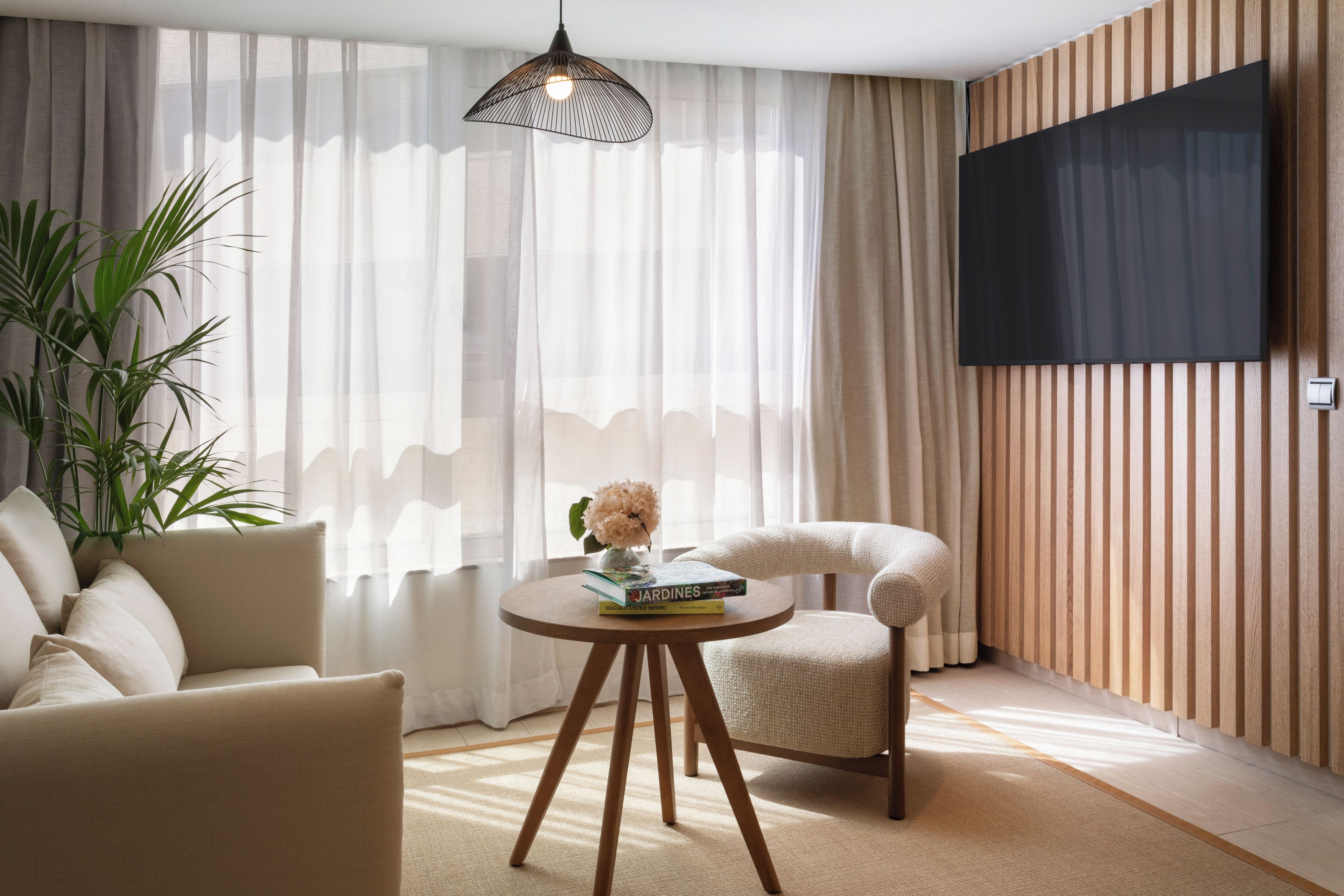 A modern living room with a sofa, a chair, a round table with a book and a vase, a floor lamp, and a TV mounted on the wall.