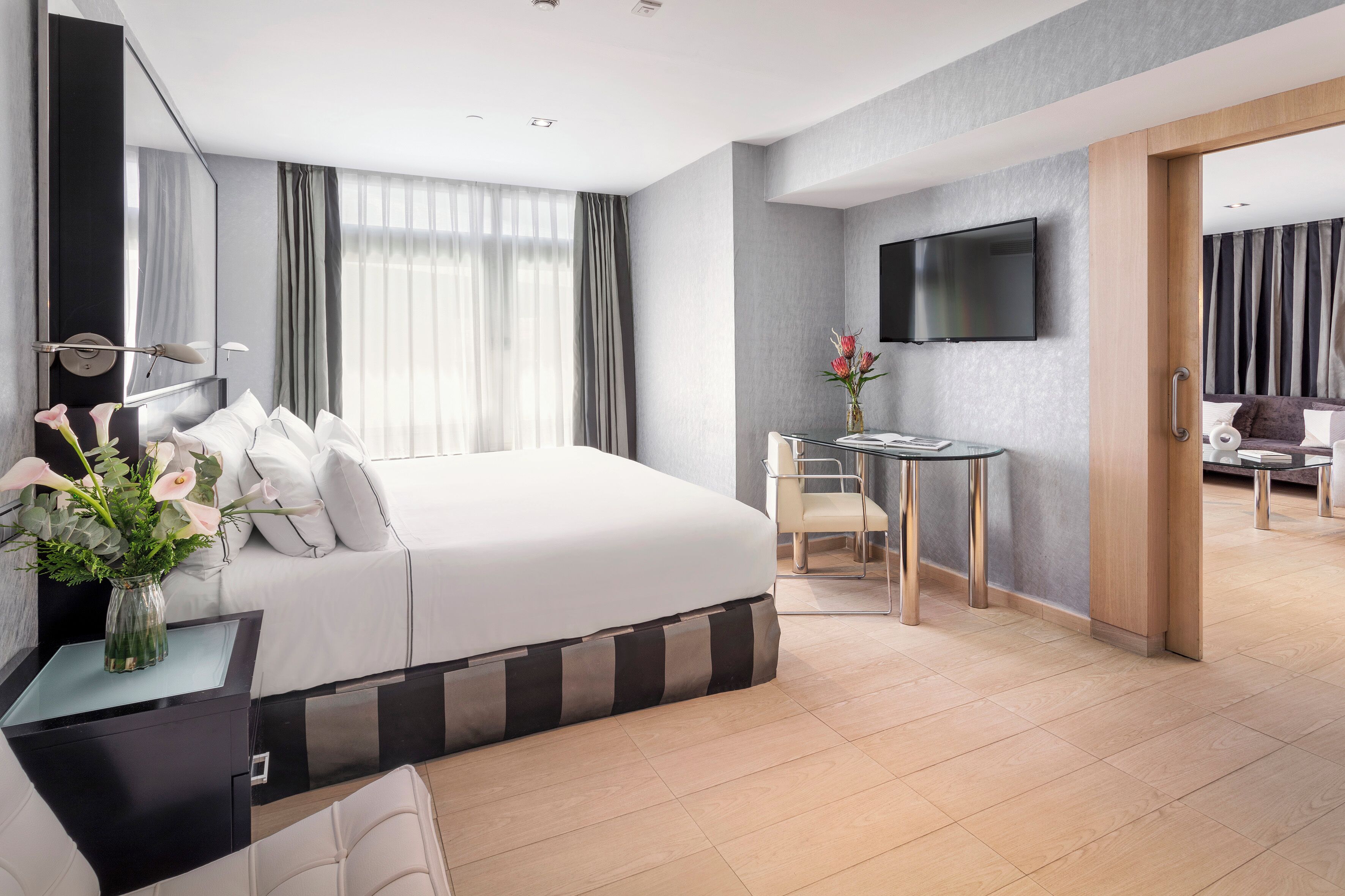 Una habitación de hotel moderna con una cama grande, una mesita de noche con un jarrón de flores, un escritorio pequeño con una silla, un televisor de pantalla plana montado en la pared y vistas de la habitación contigua.