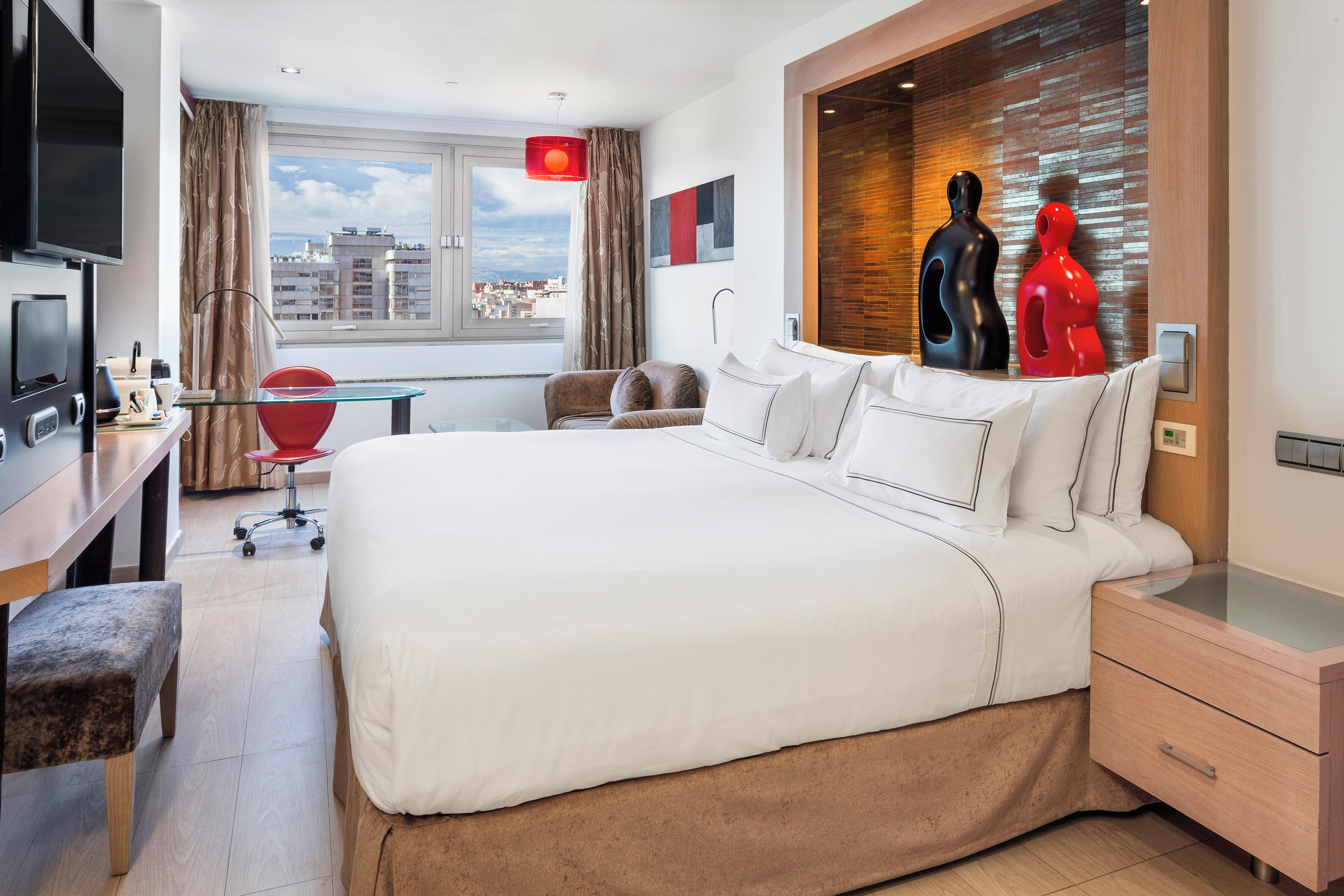 Una habitación de hotel moderna con una cama grande, un escritorio, una silla roja y vistas a la ciudad.