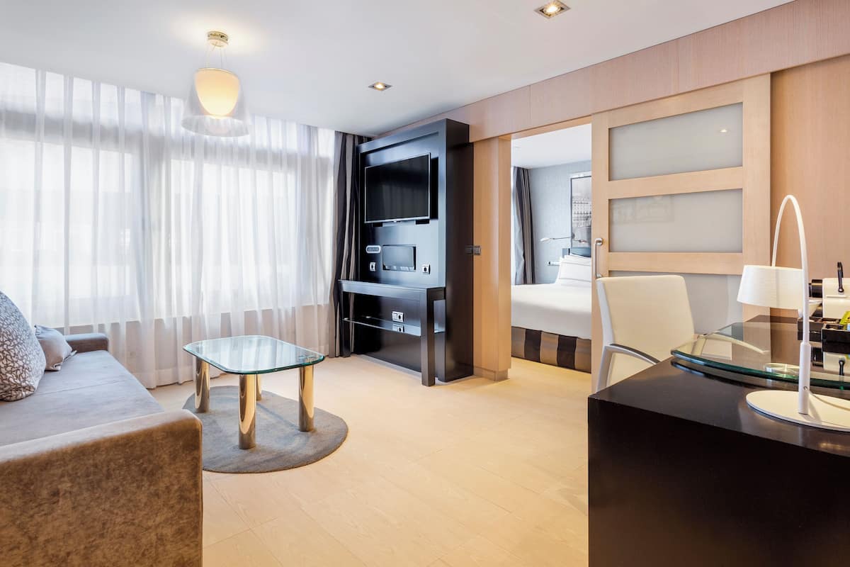 A modern hotel room with a sofa, a glass table, a TV, and a desk with a lamp.