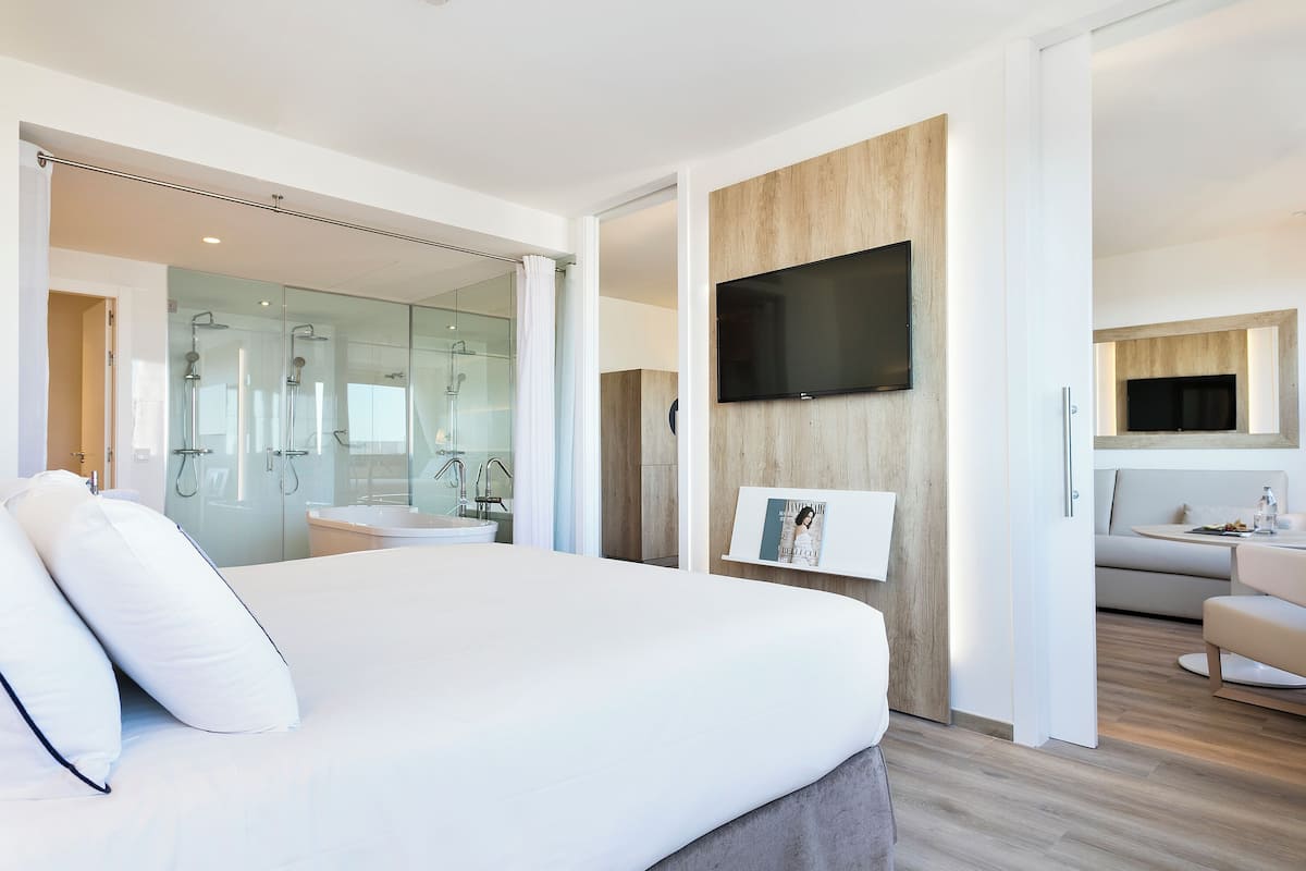 A modern hotel room with a large bed, a glass-enclosed shower area, a mounted TV, and a seating area with a sofa and table.