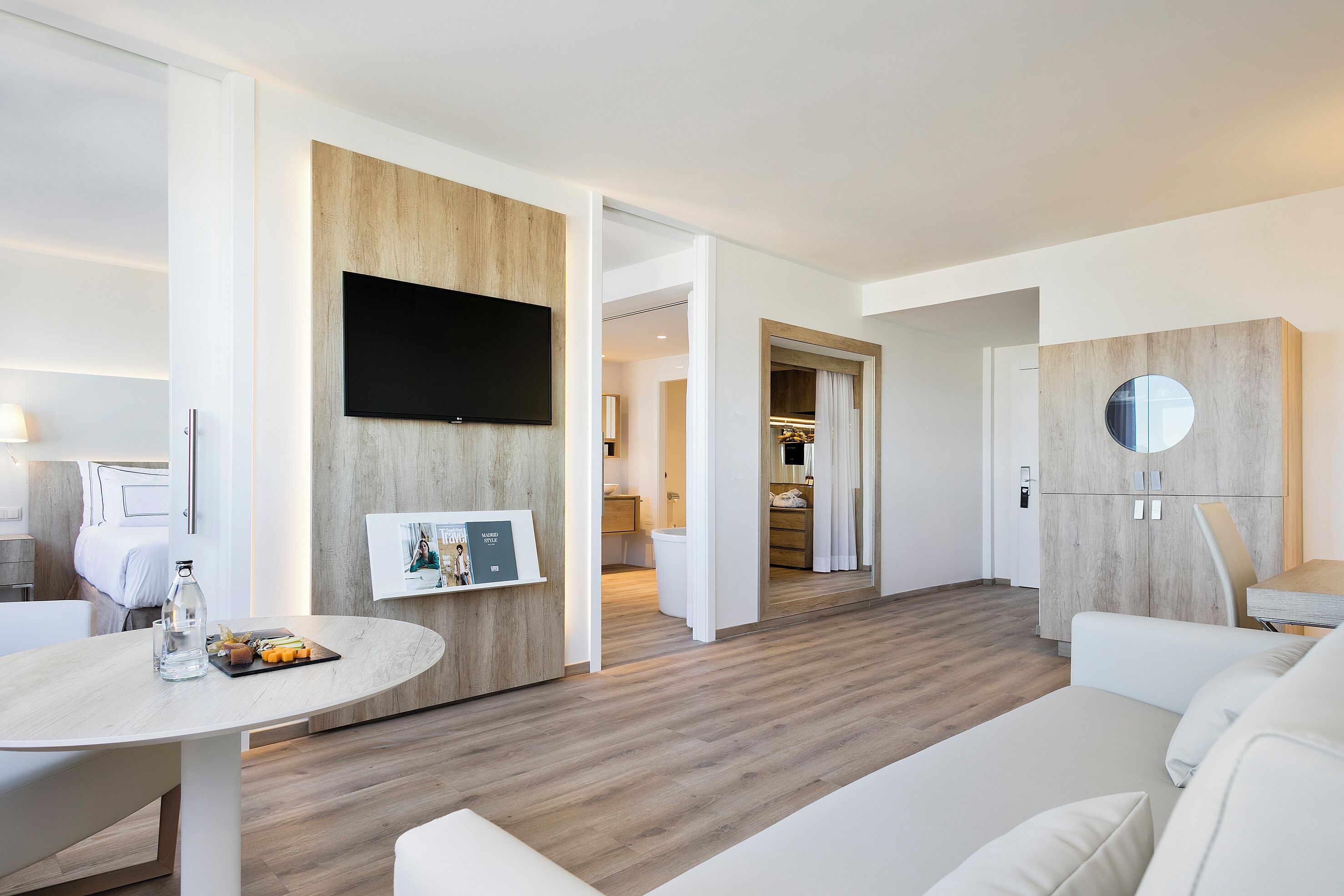 A modern hotel room with a wooden floor, a flat-screen TV mounted on a wall, a round mirror, and a dining area with a table and chairs.