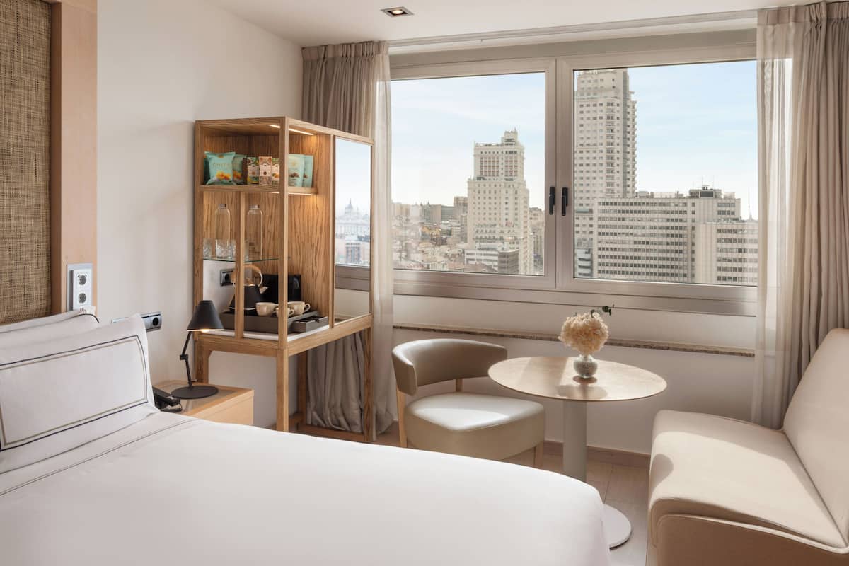 A hotel room with a bed, a seating area with a table, a bookshelf, and a large window with a city view.