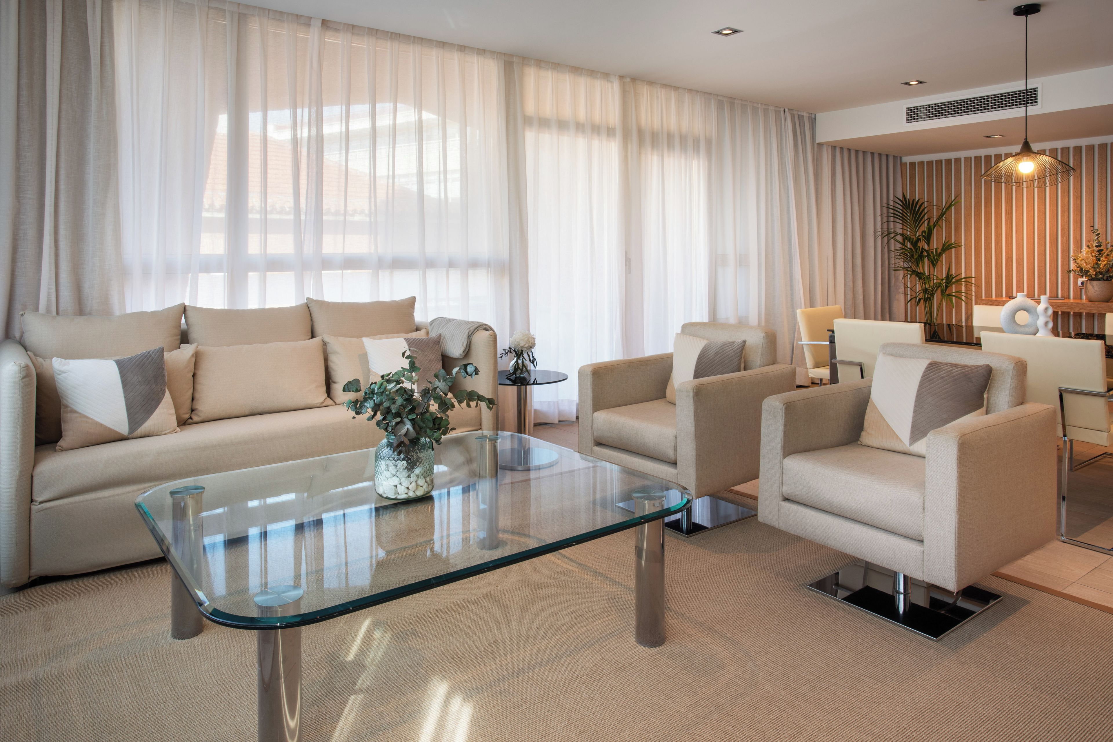 A modern living room with a glass coffee table, beige sofas, and a large window with sheer curtains.