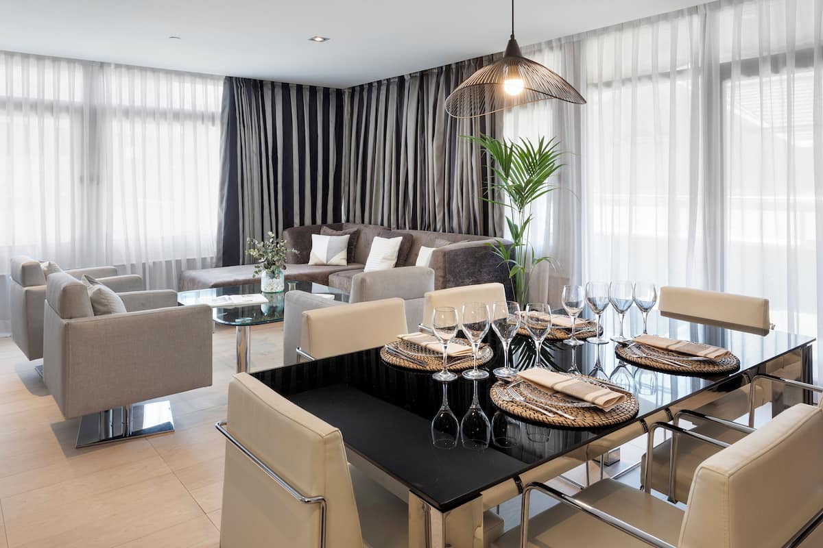 A modern living room with a glass dining table set for six, surrounded by sofas and a large window with sheer curtains.