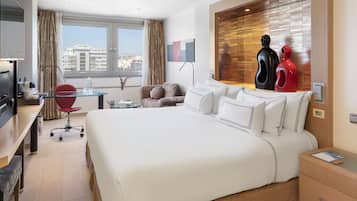 A modern hotel room with a large bed, a desk, a chair, and a view of the city.