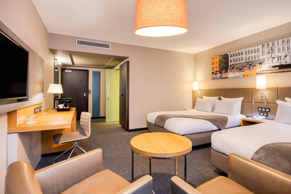 Ein Hotelzimmer mit zwei Betten, einem Schreibtisch mit Stuhl, einem runden Tisch und einem Fernseher.