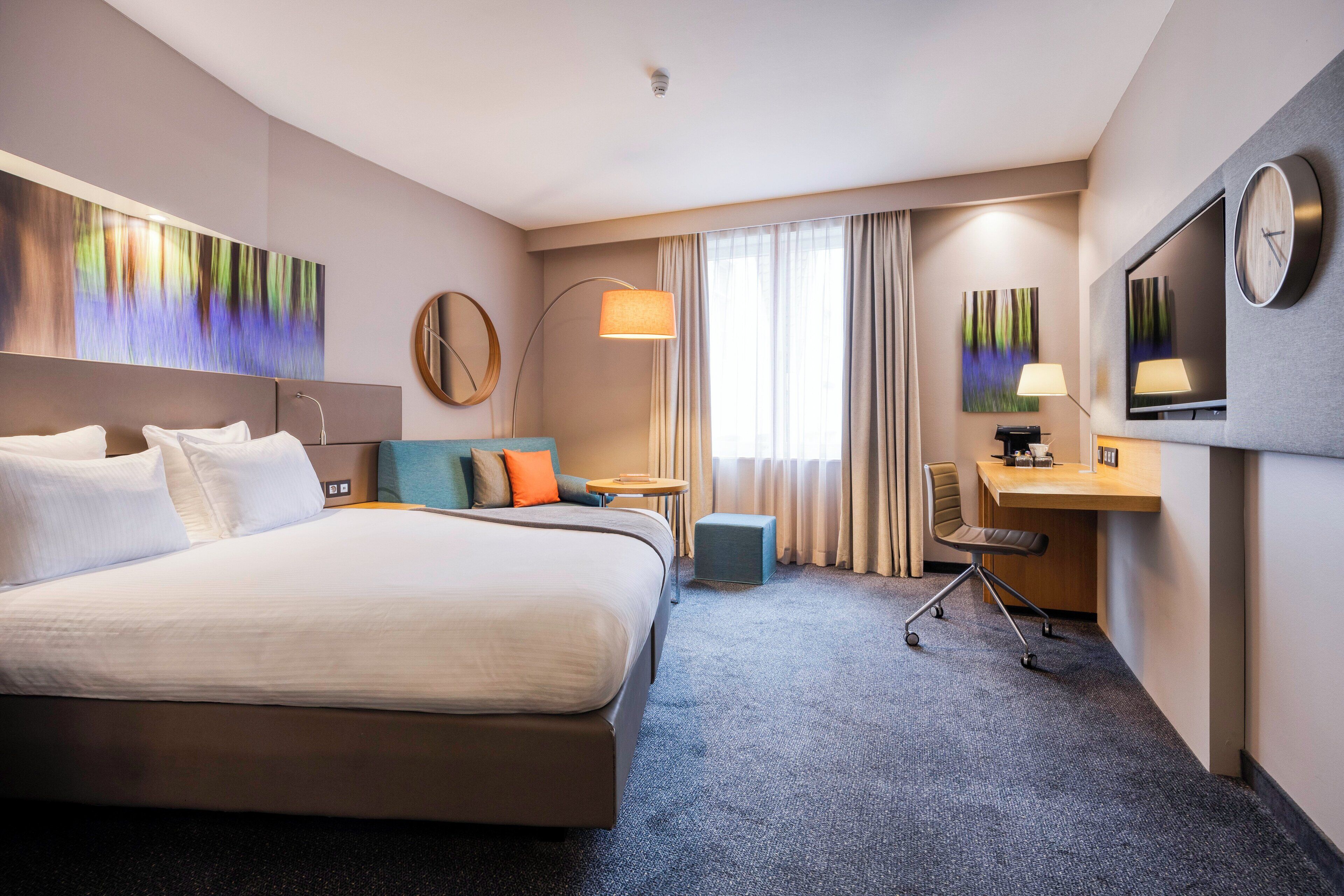 A hotel room with a large bed, a desk, a chair, a TV, and artwork on the walls.