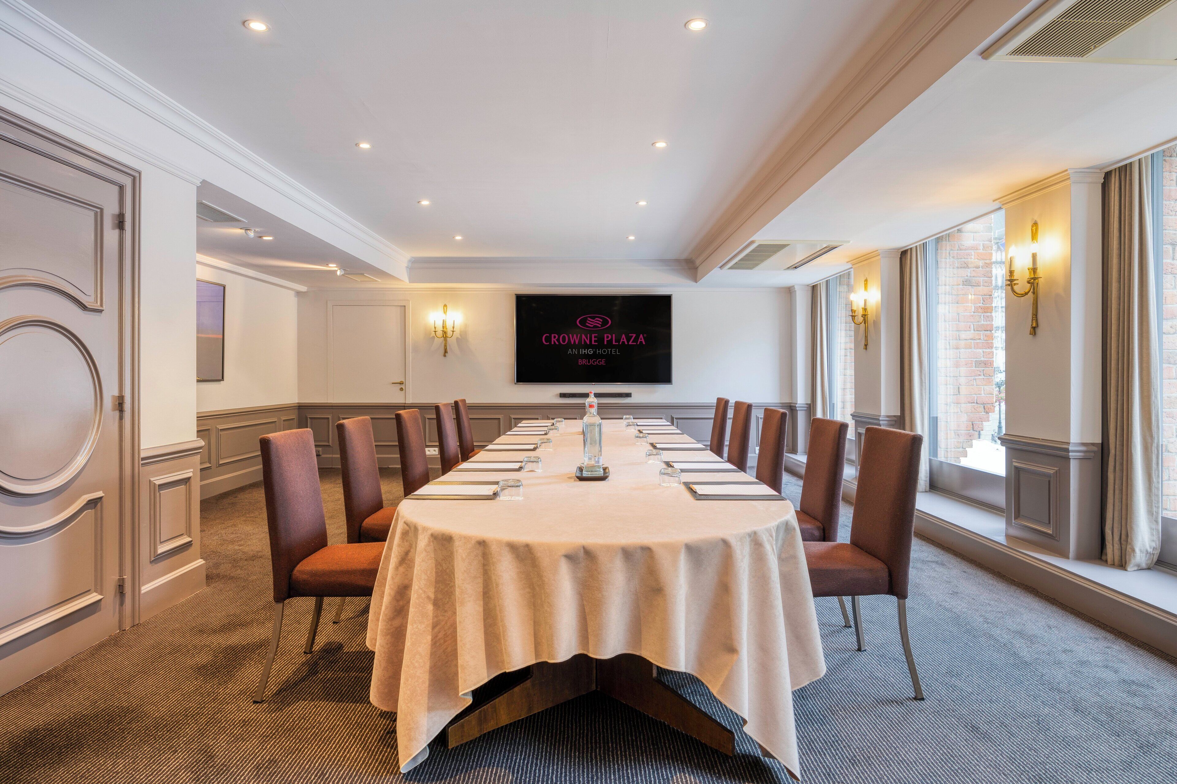 A conference room with a long table, chairs, and a television screen displaying "Crowne Plaza An ING Hotel Brugge".