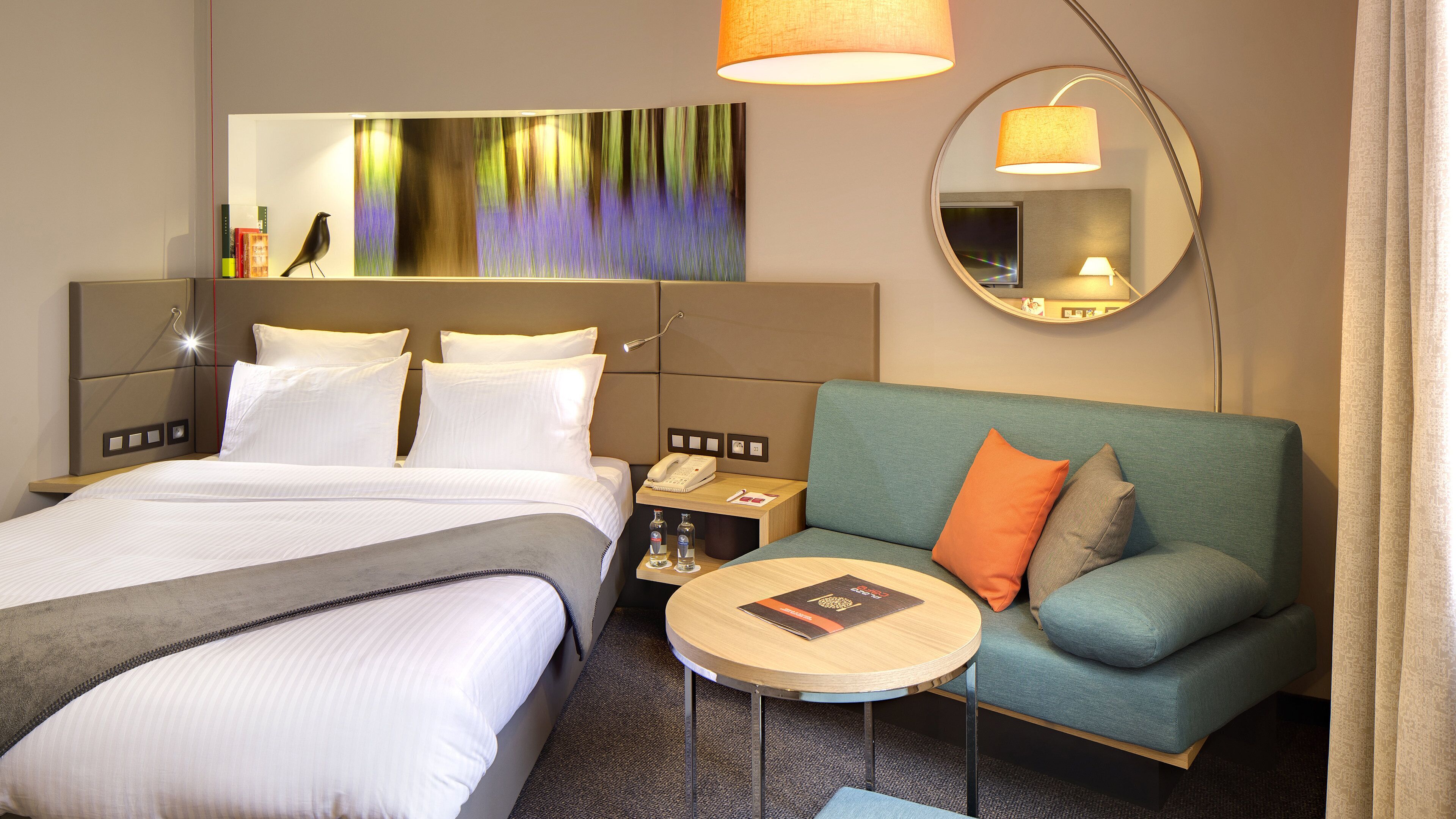 A hotel room with a bed, a sofa, a round table, a mirror, and a wall art piece.