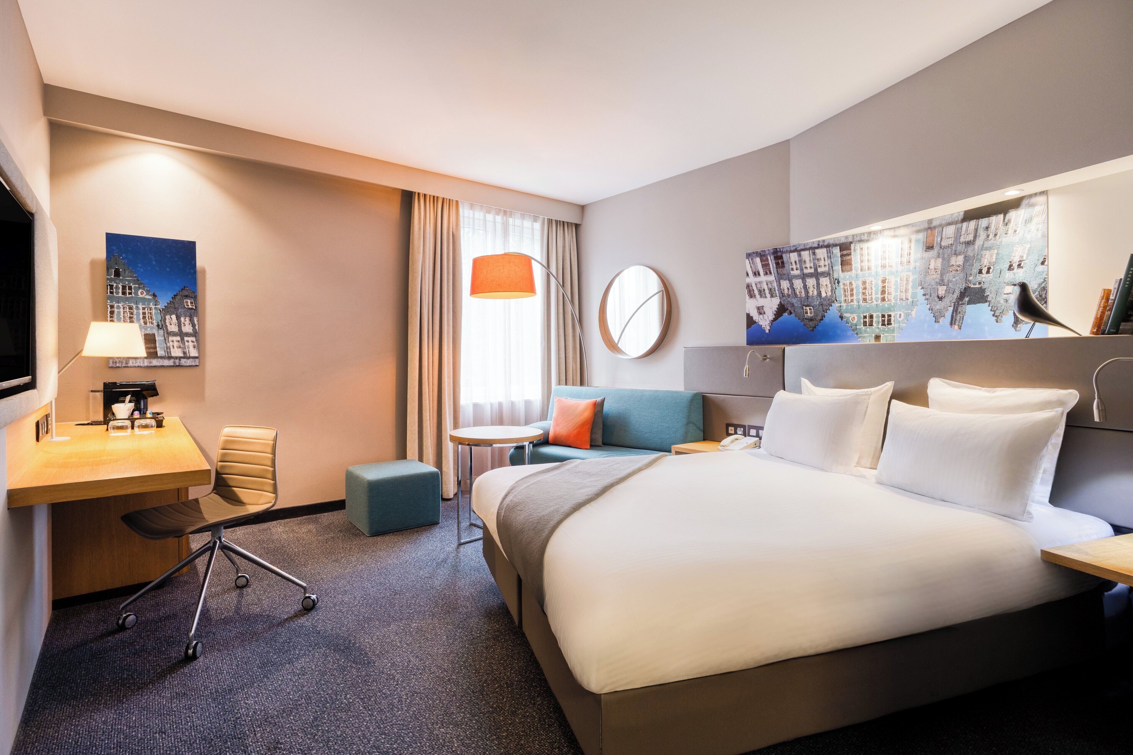 Een hotelkamer met een groot bed, een bureau met lamp, een stoel, een kleine tafel en een tv aan de muur.