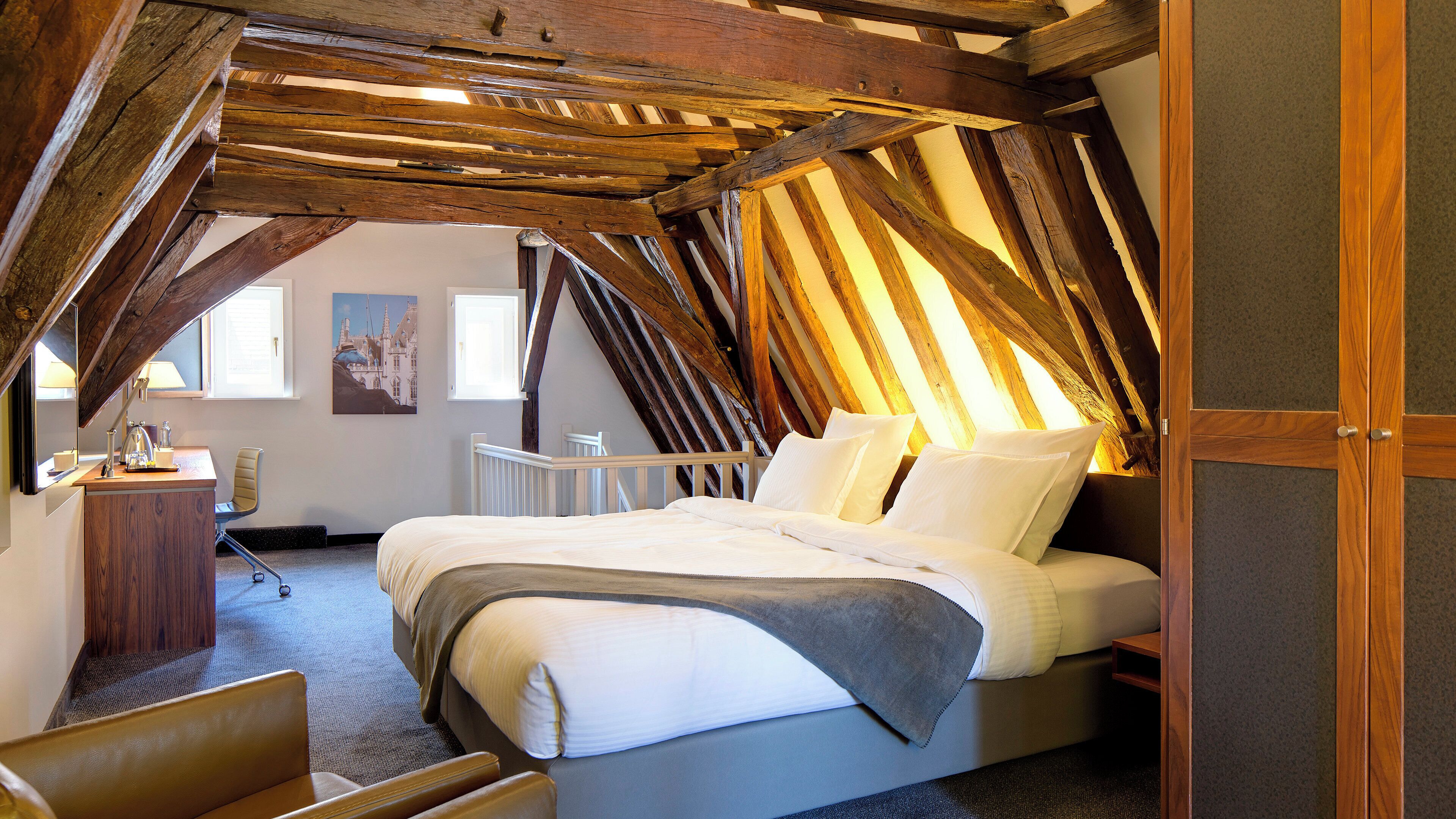 A bedroom with a sloped ceiling, a bed, a chair, and a desk with a lamp.