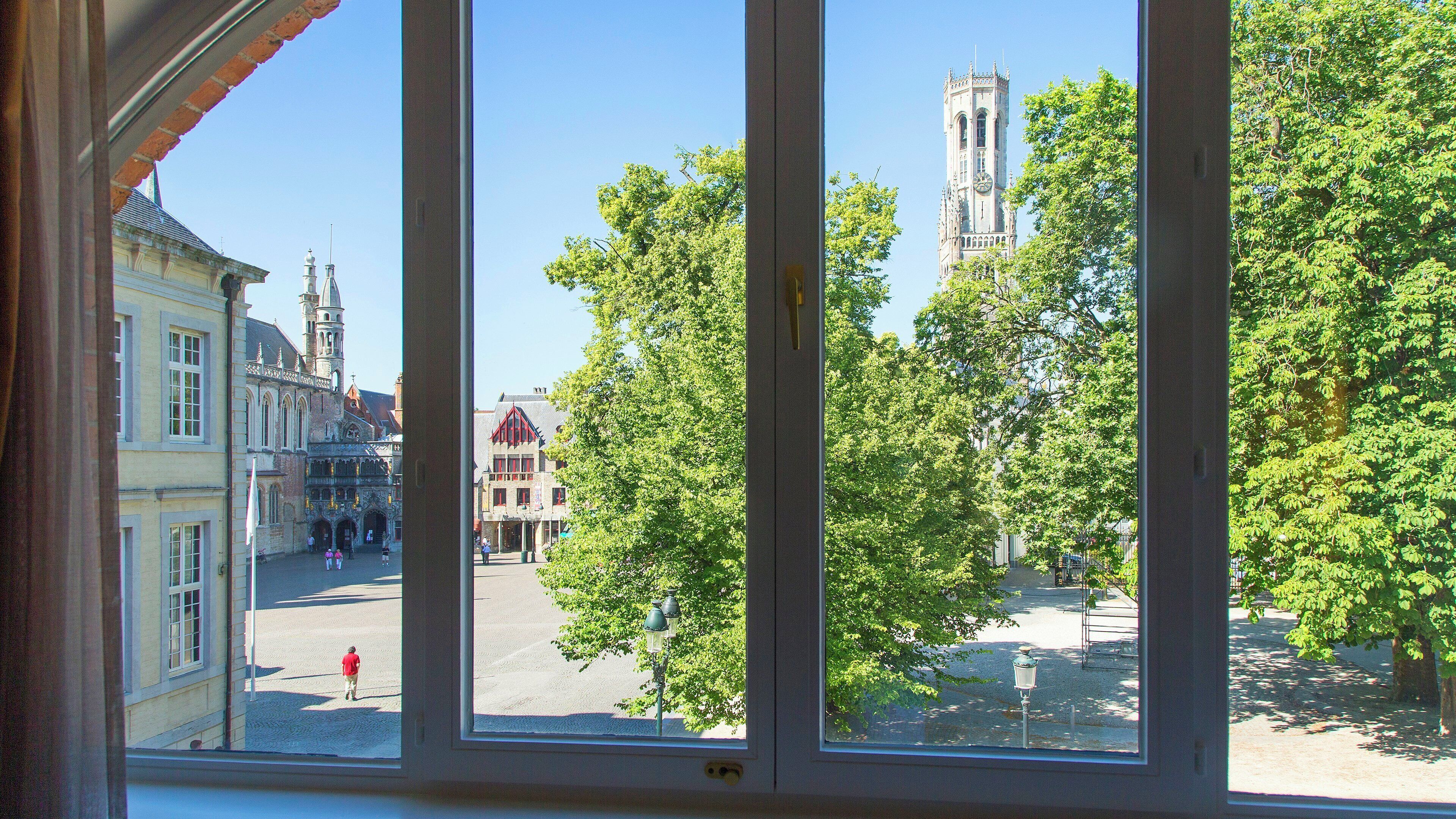 Uitzicht door een raam op een stadsplein met historische gebouwen, waaronder een kerktoren, en weelderige groene bomen.