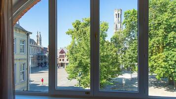 Uitzicht door een raam op een stadsplein met historische gebouwen, waaronder een kerktoren, en weelderige groene bomen.