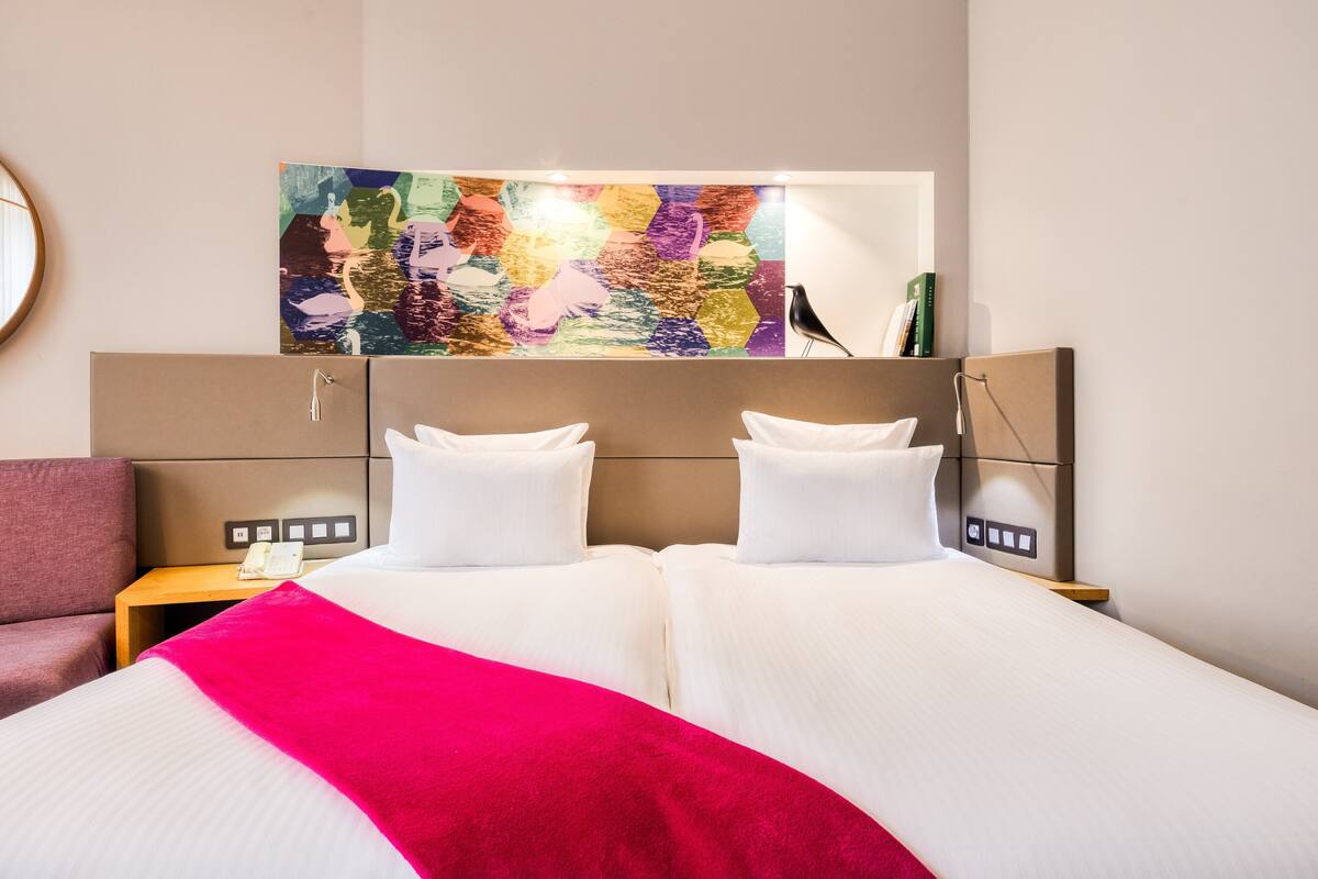 Ein Hotelzimmer mit zwei Betten, einem farbenfrohen abstrakten Gemälde darüber, einem Nachttisch mit Telefon und einem Sessel.