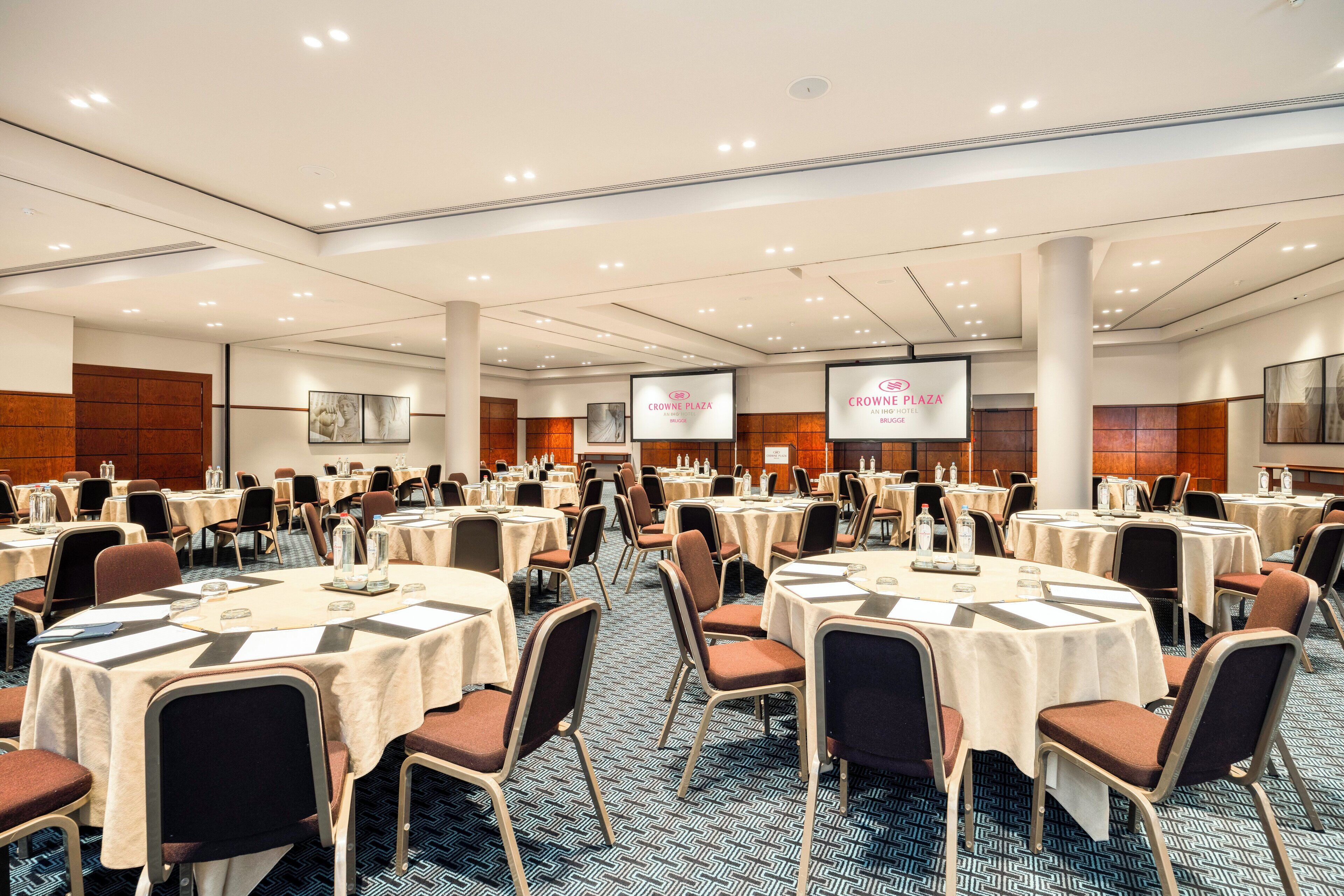 Een ruime congreszaal met meerdere ronde tafels, stoelen en een scherm waarop “CROWNE PLAZA” staat.