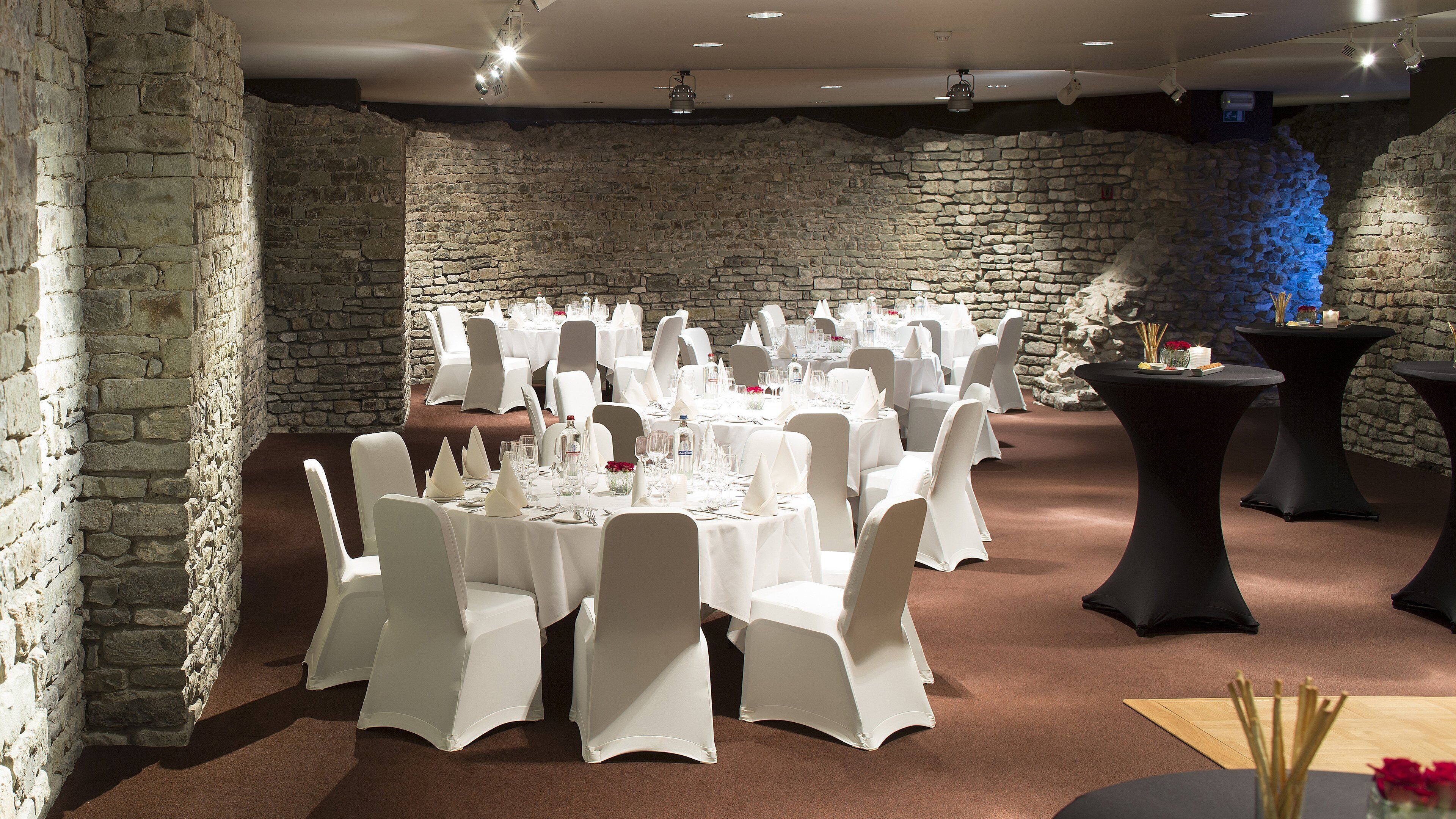 Een feestzaal met stenen muren, ronde tafels en witte stoelen.