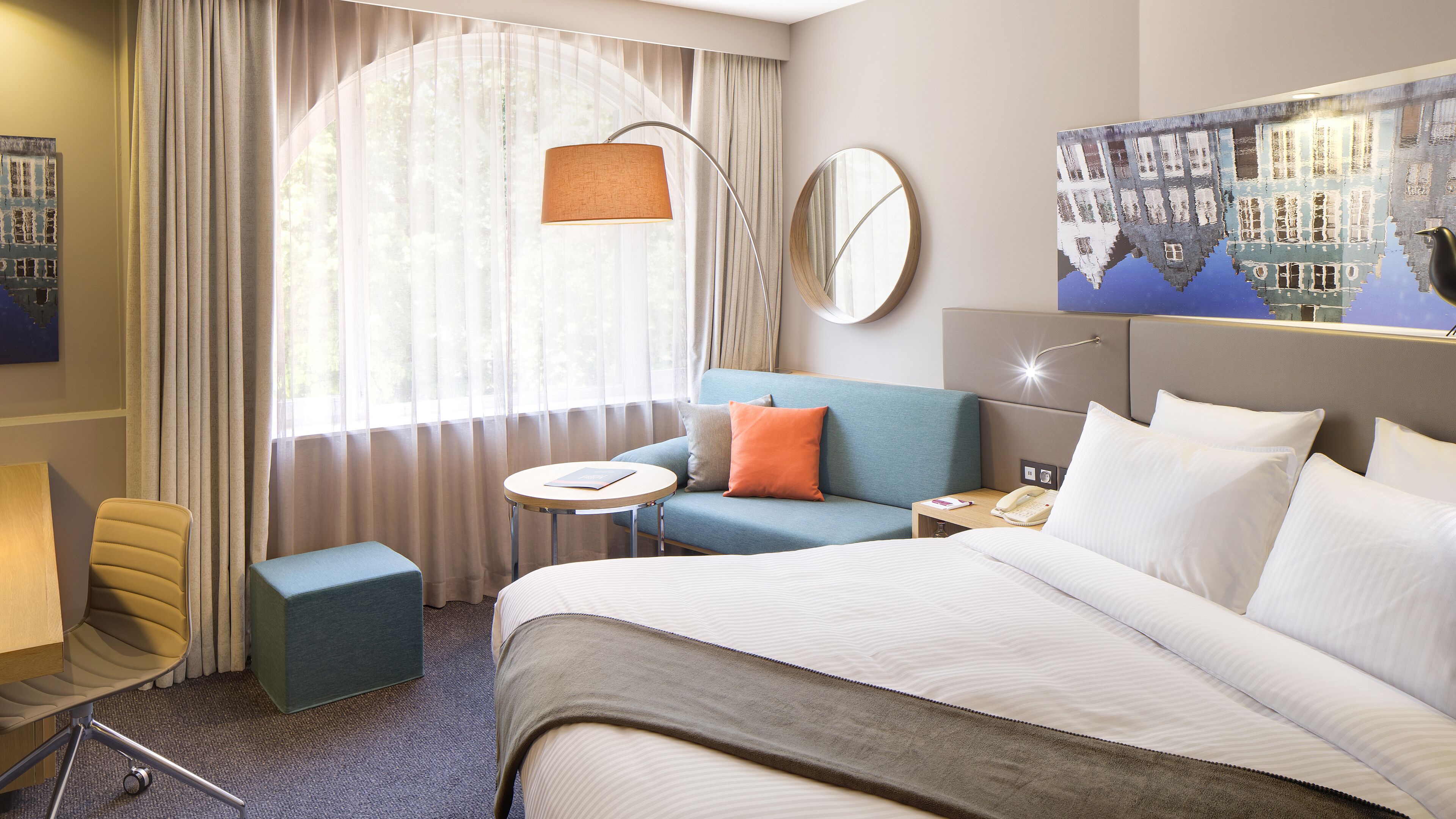 A hotel room with a bed, a sofa, a small table, a chair, a desk, a lamp, a mirror, and a painting on the wall.