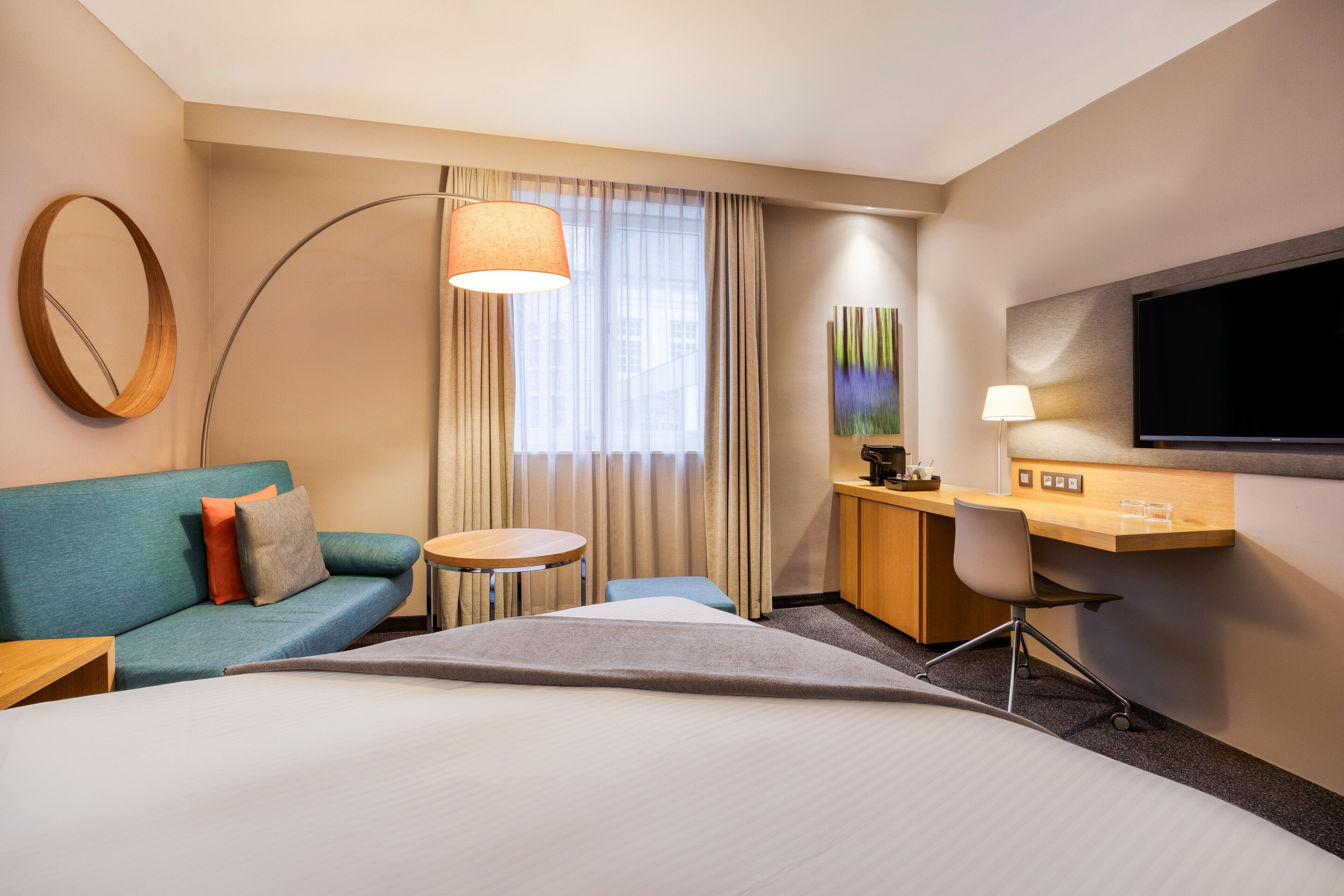 Habitación de hotel con una cama grande, un sofá, un escritorio con silla, televisor y un amplio ventanal con cortinas.