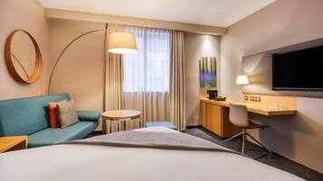Habitación de hotel con una cama grande, un sofá, un escritorio con silla, televisor y un amplio ventanal con cortinas.