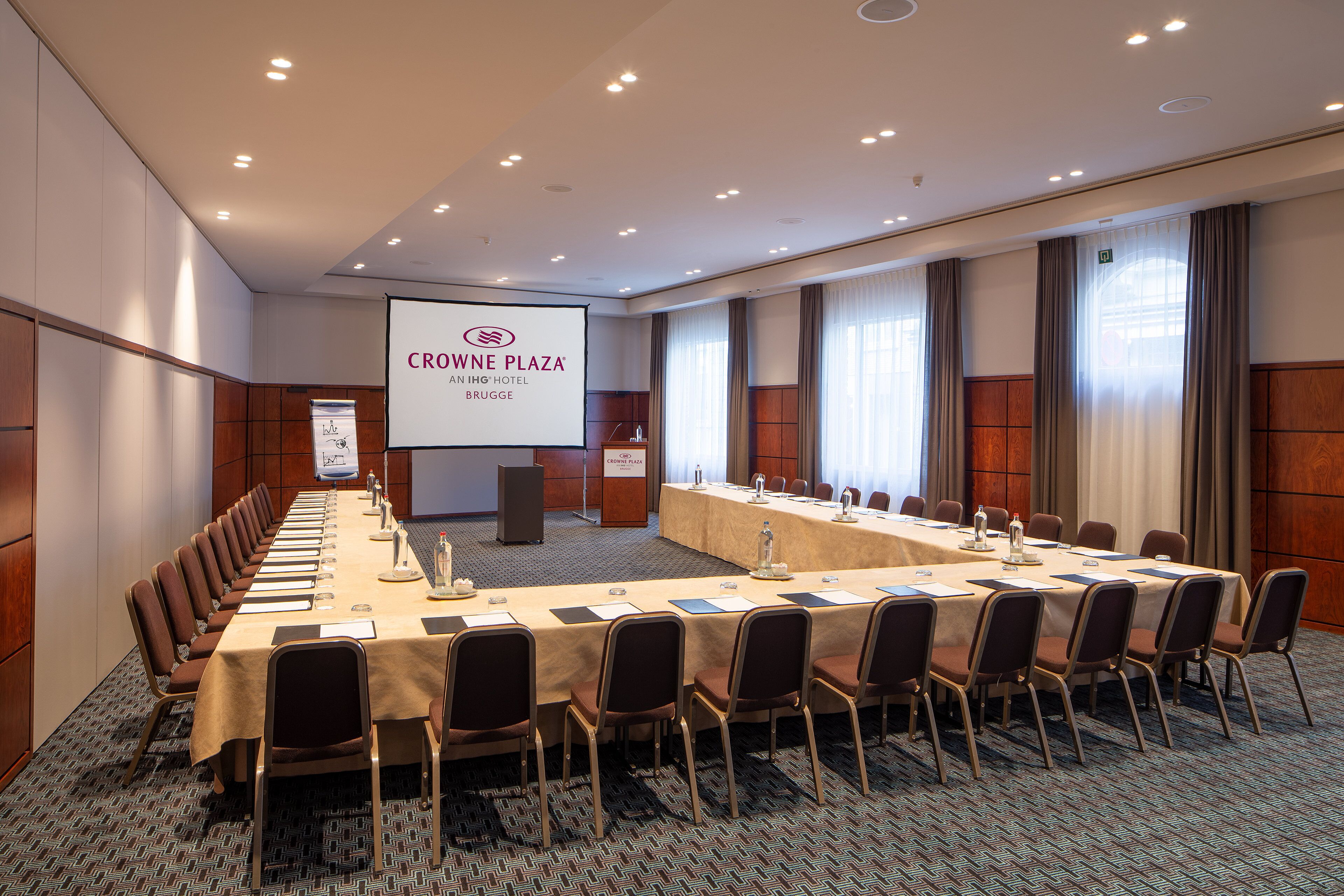 Un salón de conferencias con una larga mesa, sillas y una pantalla que muestra el logotipo de Crohne Plaza.