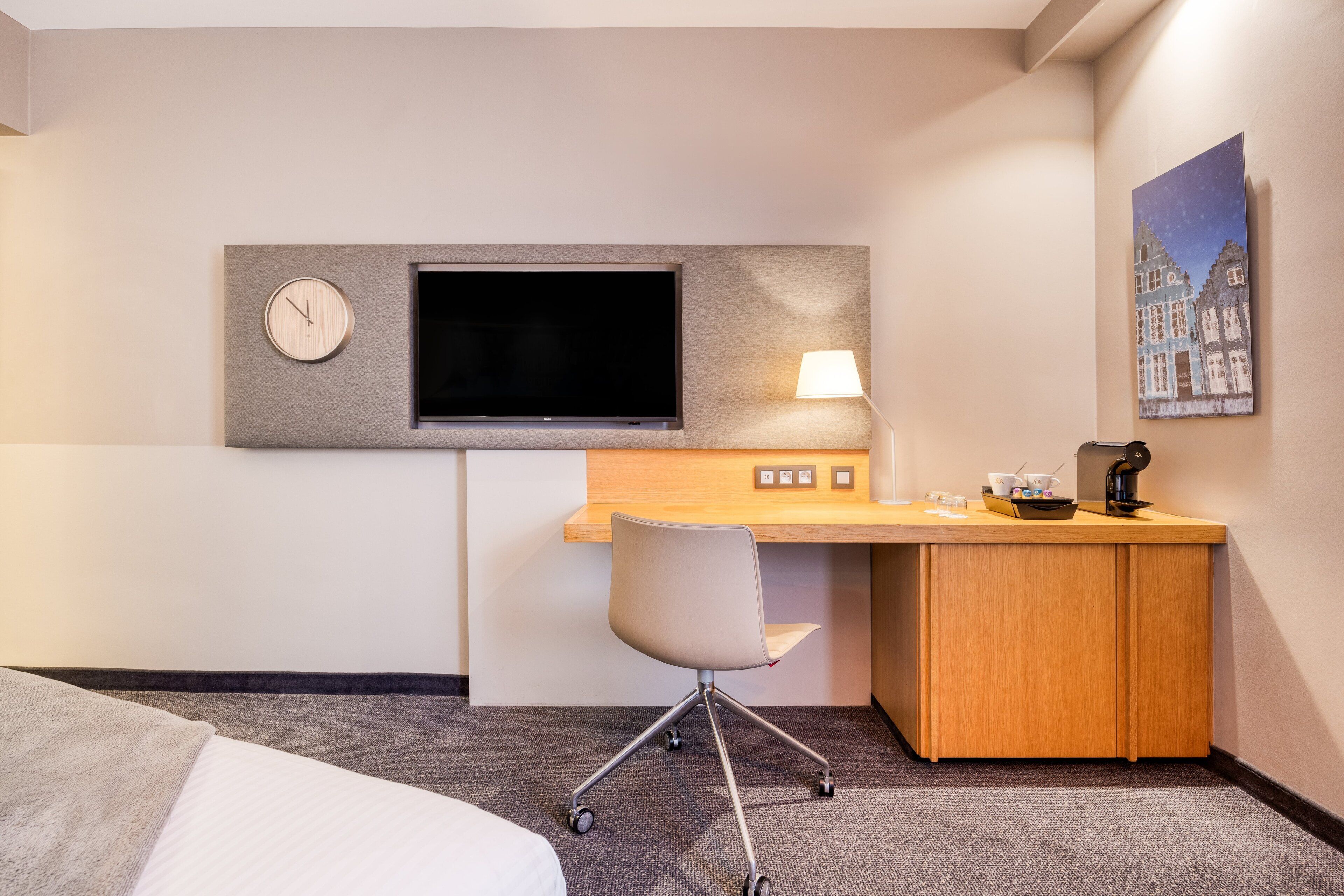 A hotel room with a bed, a desk with a chair, a flat-screen TV, a lamp, and a painting on the wall.