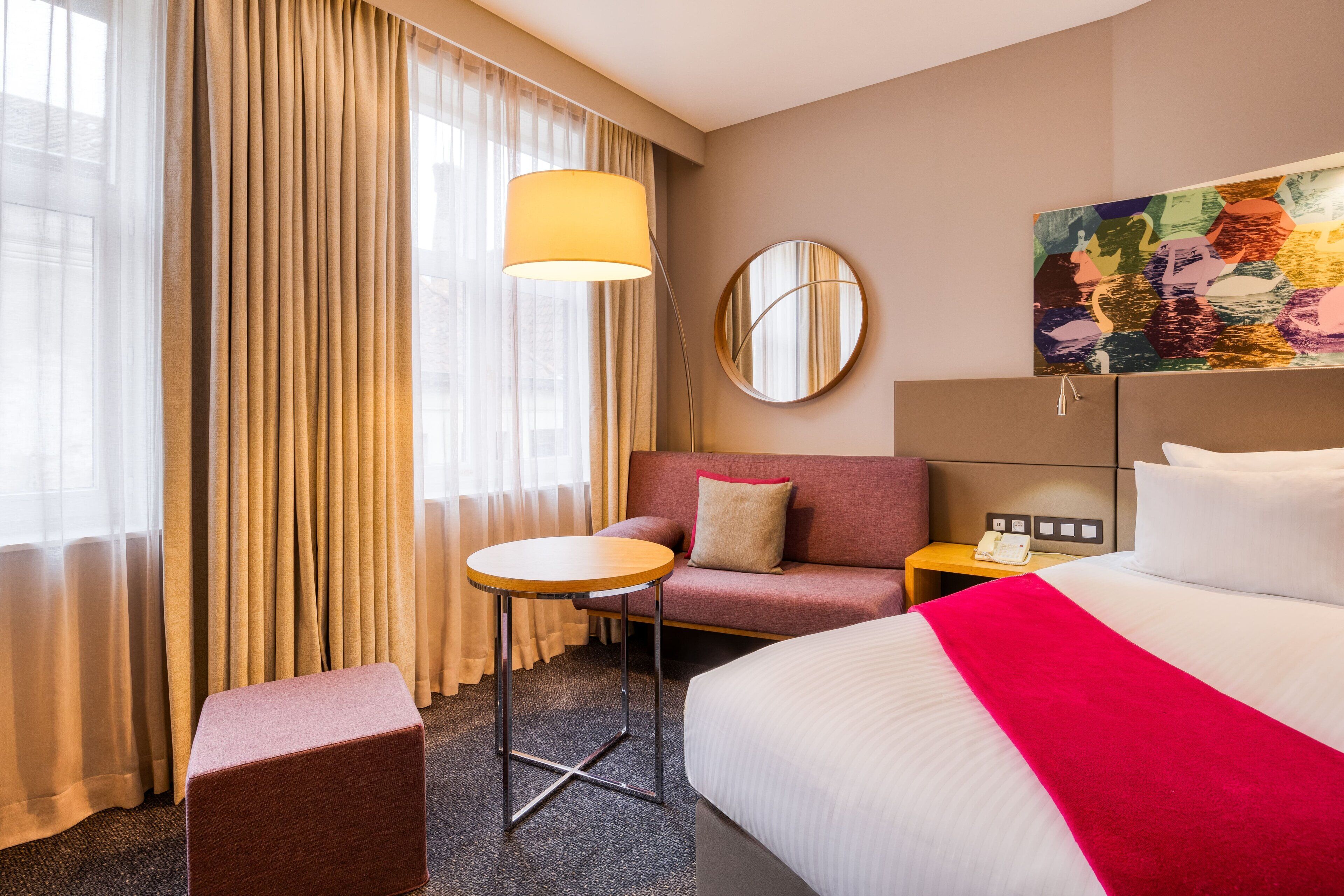 A hotel room with a bed, a round mirror, a small table, a sofa, and a painting on the wall.