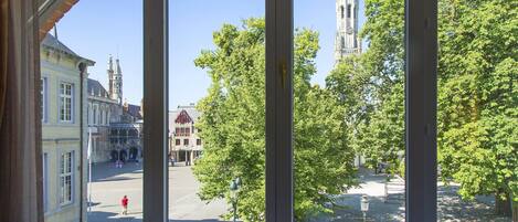 Vista desde una ventana de una plaza de pueblo con edificios históricos, incluyendo una torre de iglesia, y frondosos árboles verdes.