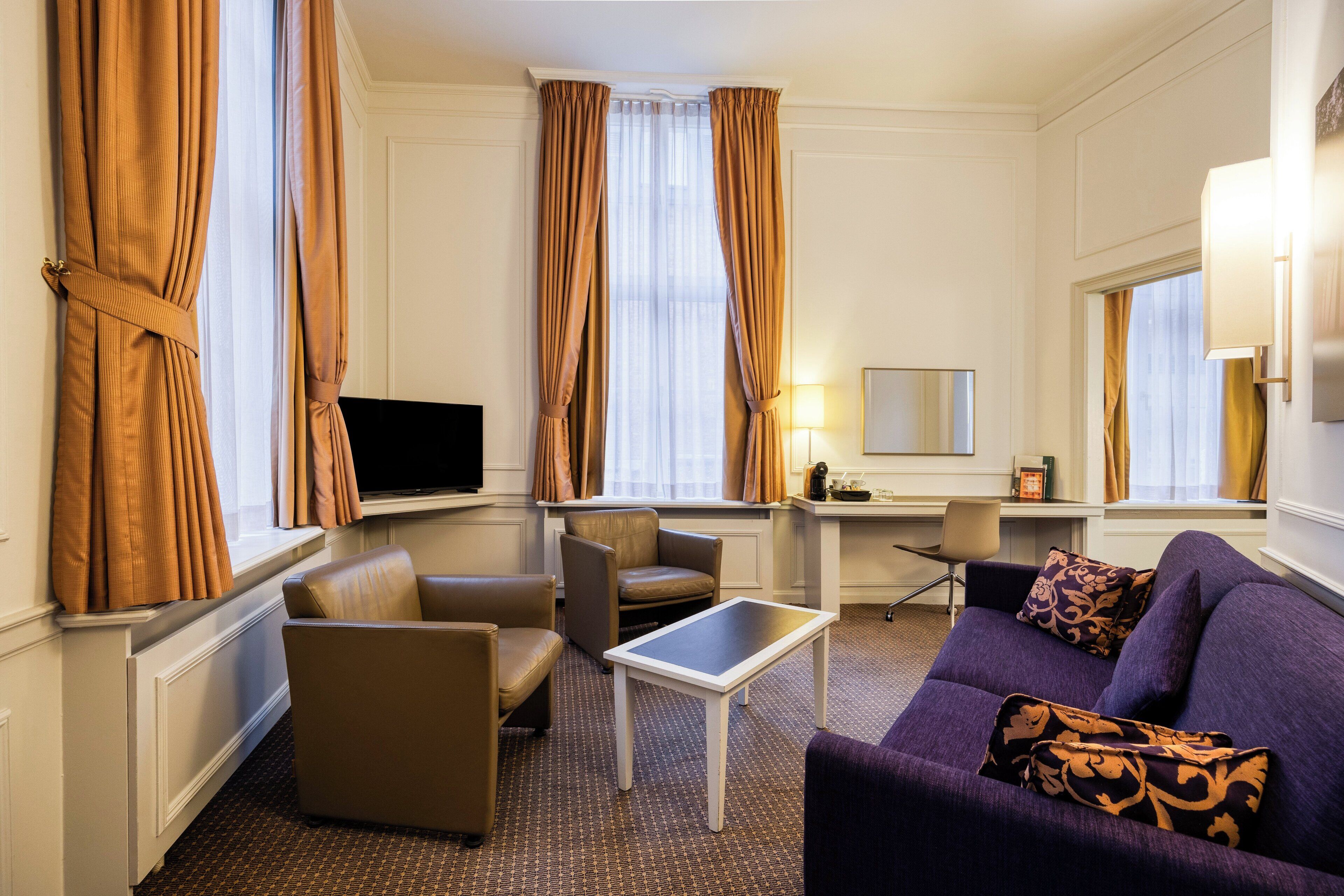 A modern hotel room with a sofa, armchairs, a small table, a TV, and a desk with a chair.