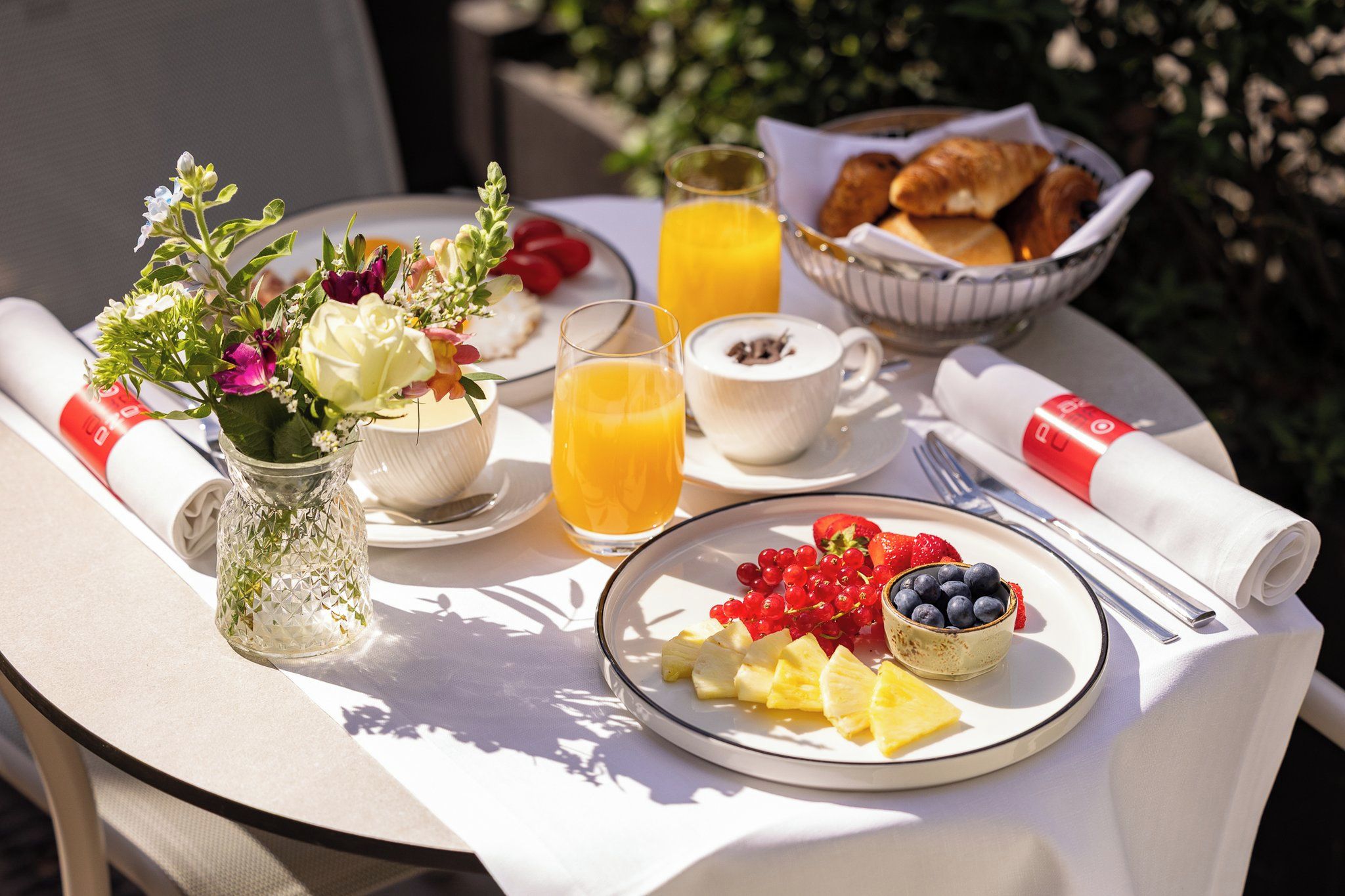Una mesa de desayuno con un plato de fruta, un vaso de jugo de naranja, un bol de croissants y un jarrón de flores.