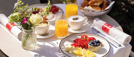 Una mesa de desayuno con un plato de fruta, un vaso de jugo de naranja, un bol de croissants y un jarrón de flores.