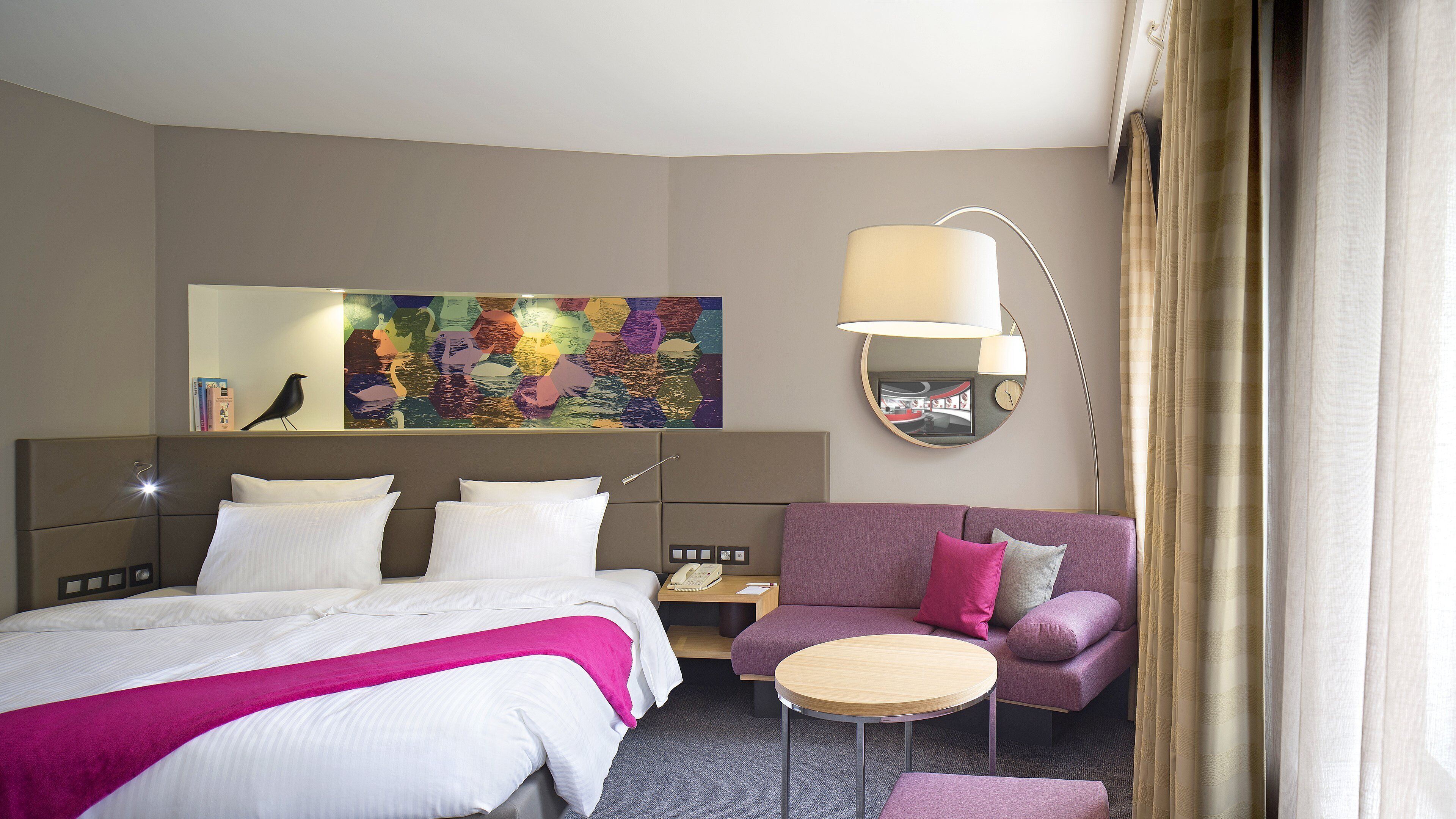A hotel room with a bed, a sofa, a small table, a lamp, a clock, and a colorful abstract painting on the wall.