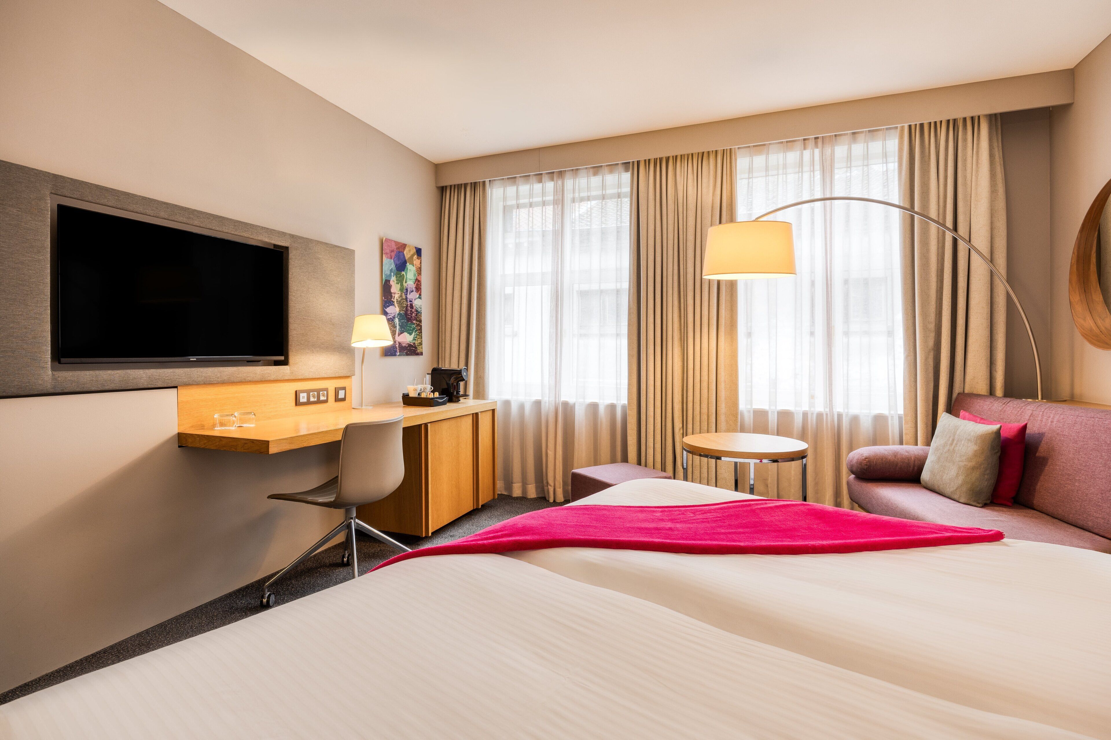 A hotel room with a bed, desk, chair, TV, lamp, and a sofa.