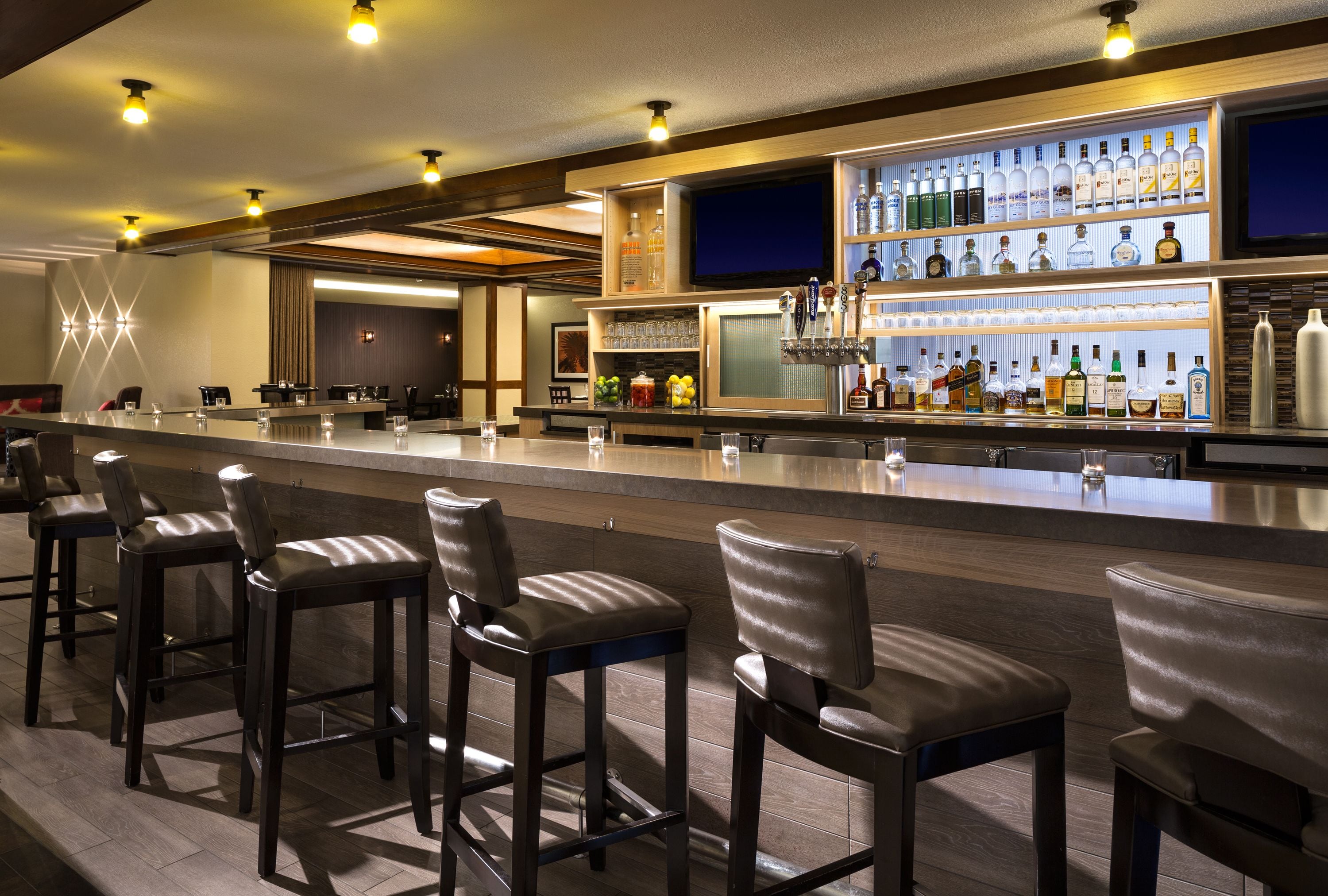 Una zona de bar con una barra larga, varios taburetes y una estantería bien surtida con diferentes botellas y vasos.