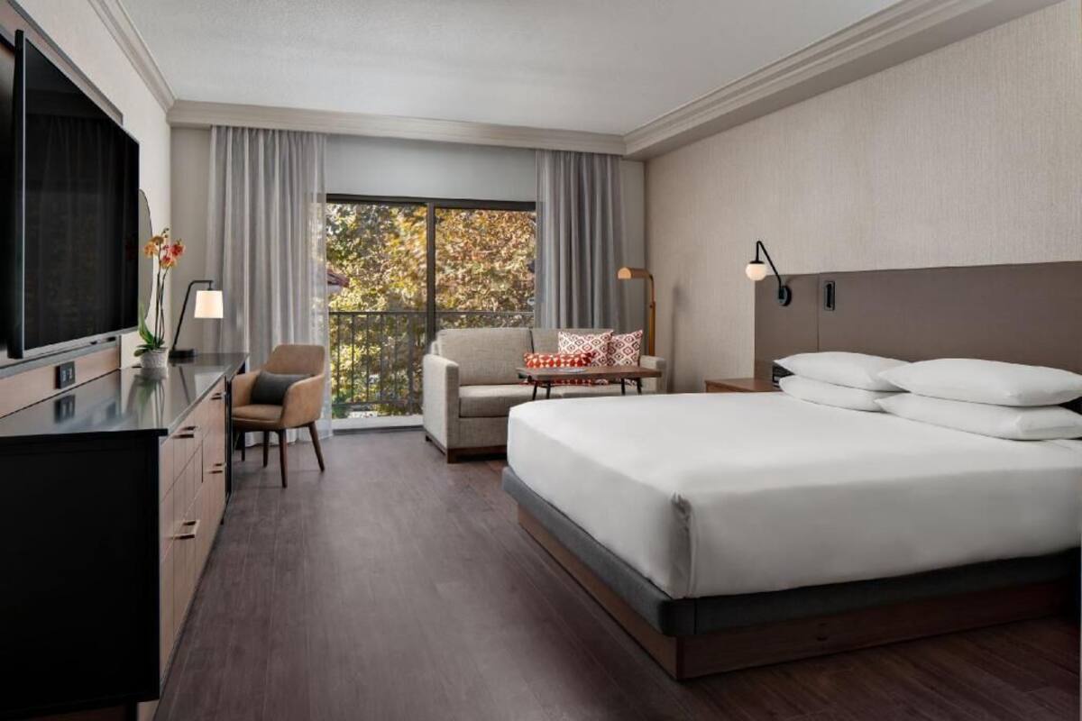 A hotel room with a bed, a desk, a chair, a small table, a TV, and a window with curtains.