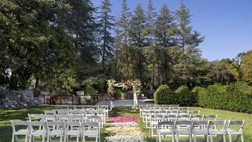 An outdoor wedding venue with white chairs arranged in rows, a floral arch, and a stone wall in the background.