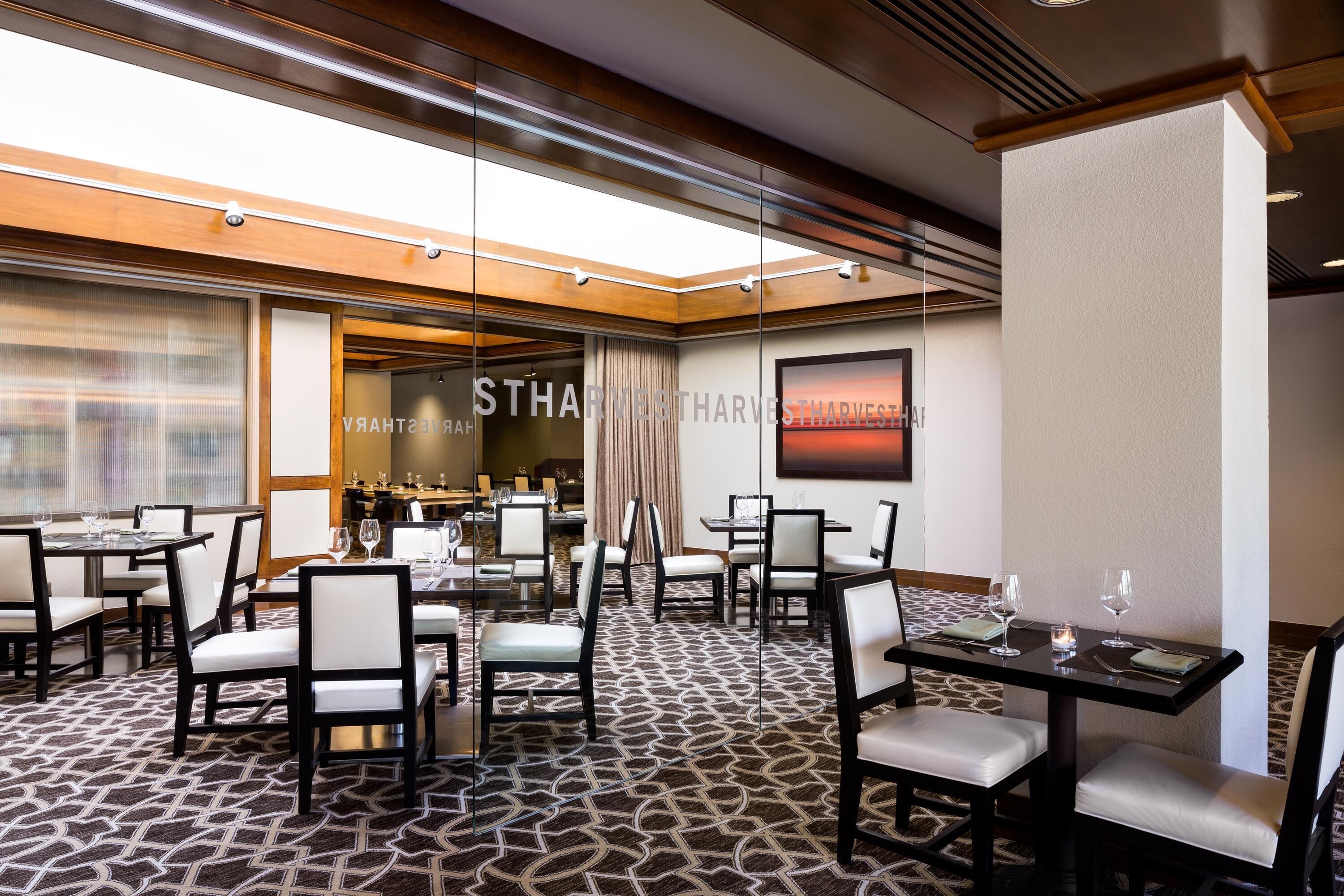 A hotel dining area with multiple tables and chairs, a patterned carpet, and a large window with curtains.