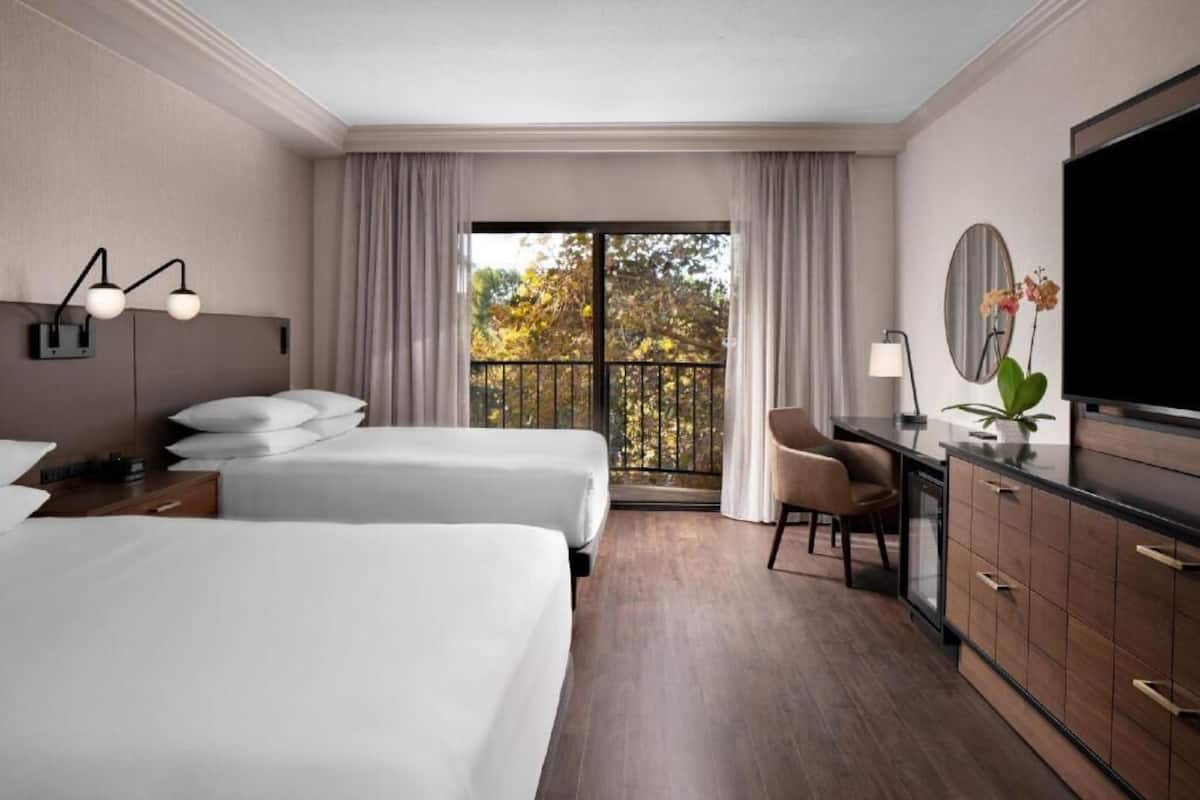Habitación de hotel con dos camas, un escritorio, un televisor, una silla y un balcón con cortinas.
