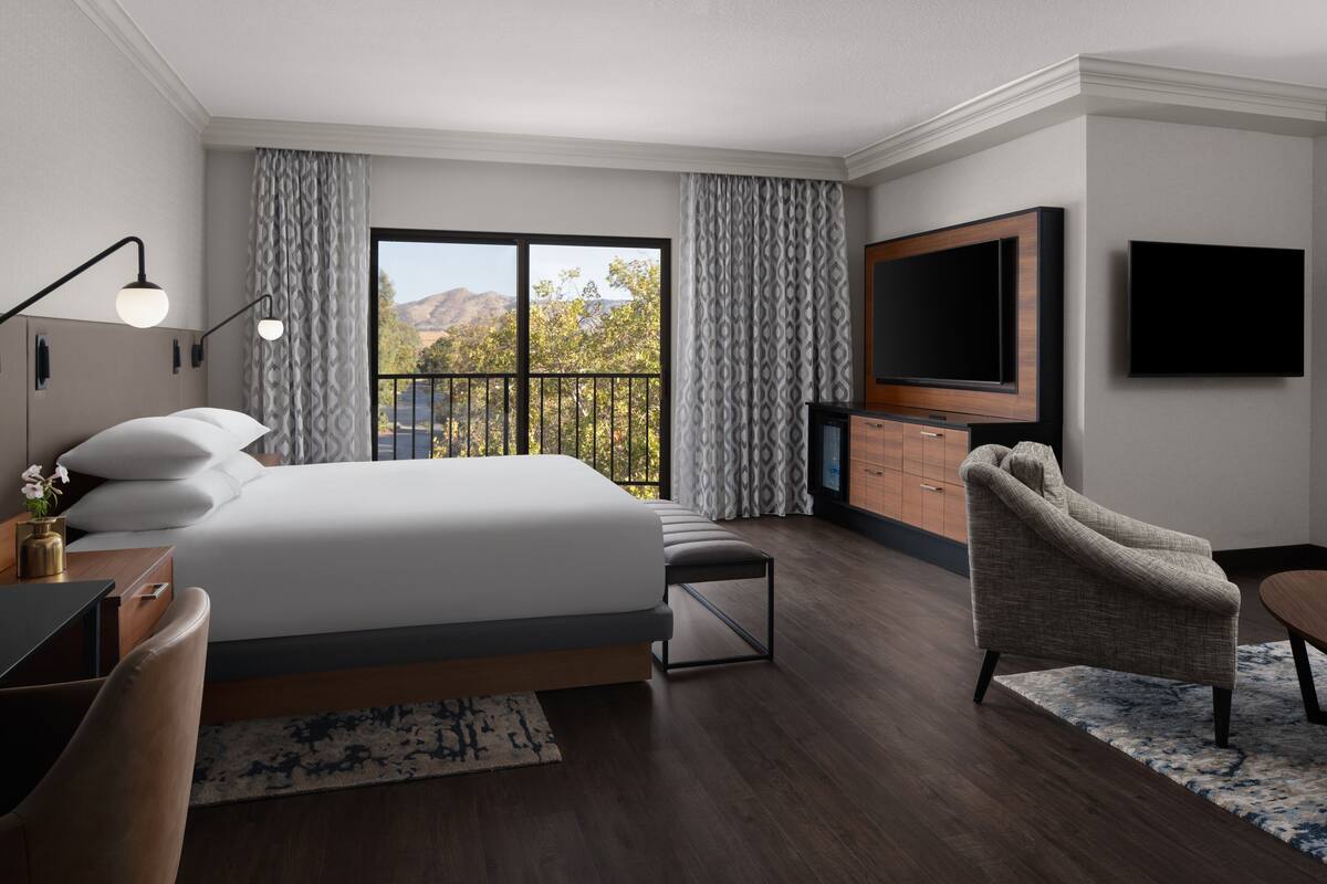 A modern hotel room with a large bed, a TV, a desk, and a chair.