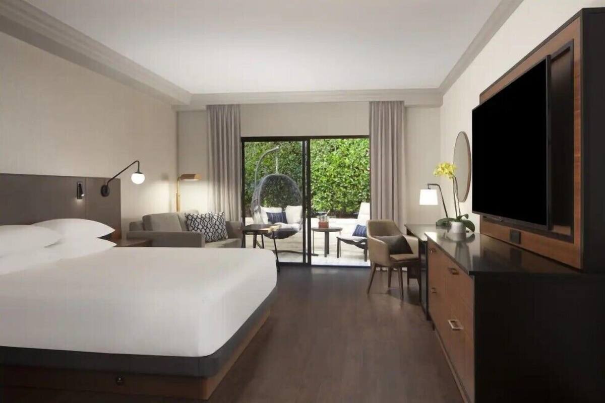 A hotel room with a large bed, a TV, a desk with a chair, and a balcony with seating.