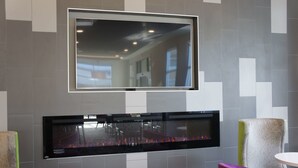 A modern interior with a flat-screen TV mounted above a fireplace, grey and white tiled walls, and purple chairs. - La Quinta Inn & Suites by Wyndham Mobile (Mobile)