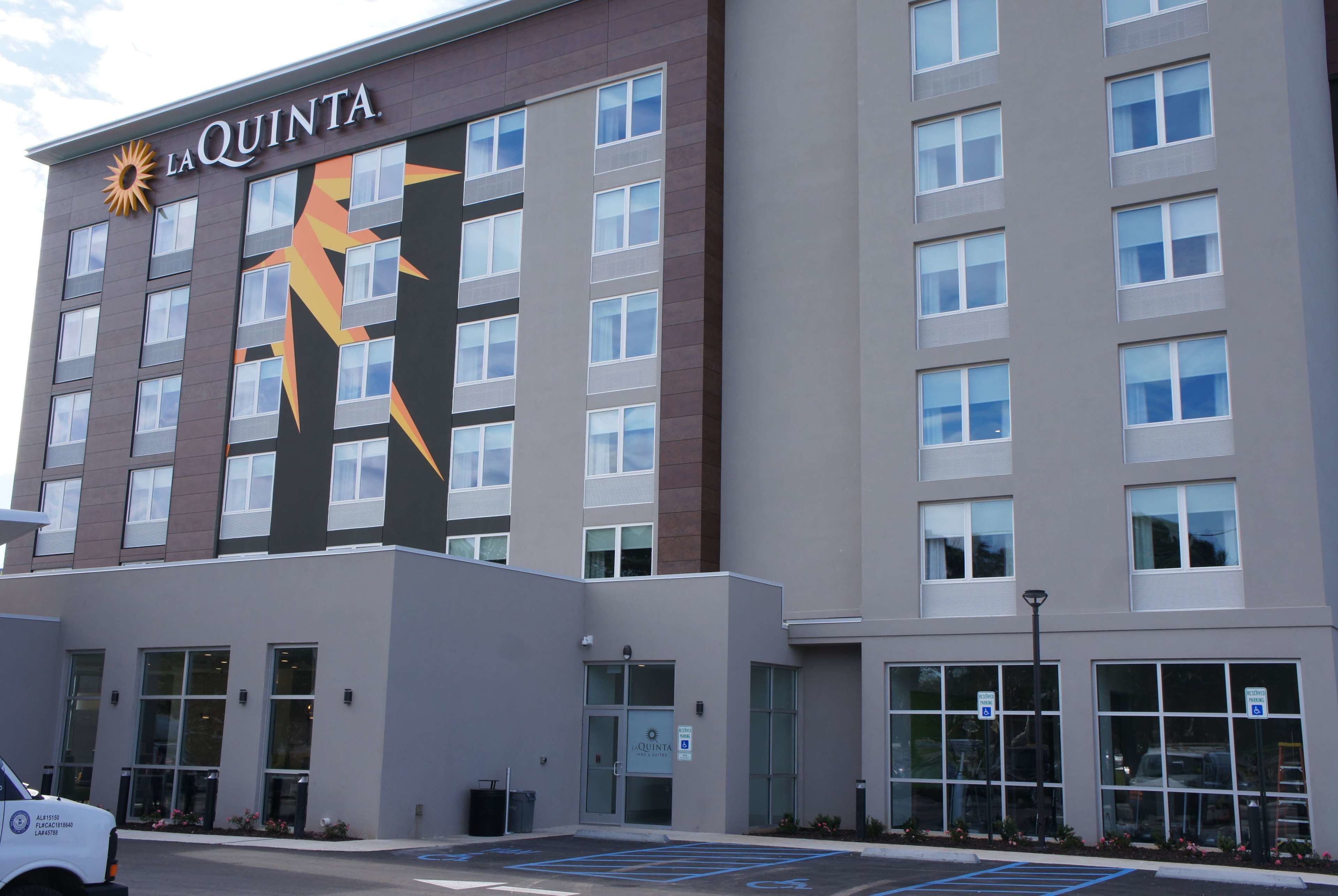A multi-story hotel with a prominent logo on the facade, large windows, and a parking lot with marked spaces.