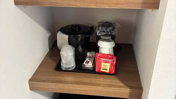 A kitchen area with a microwave on the upper shelf, a coffee maker, a kettle, and a coffee pod dispenser on the middle shelf, and a black appliance on the lower shelf.